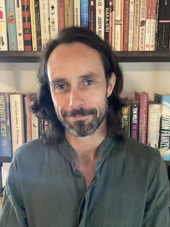 A man with shoulder-length dark hair and a beard is standing in front of a bookshelf filled with various books. He is wearing a green shirt and smiling slightly.