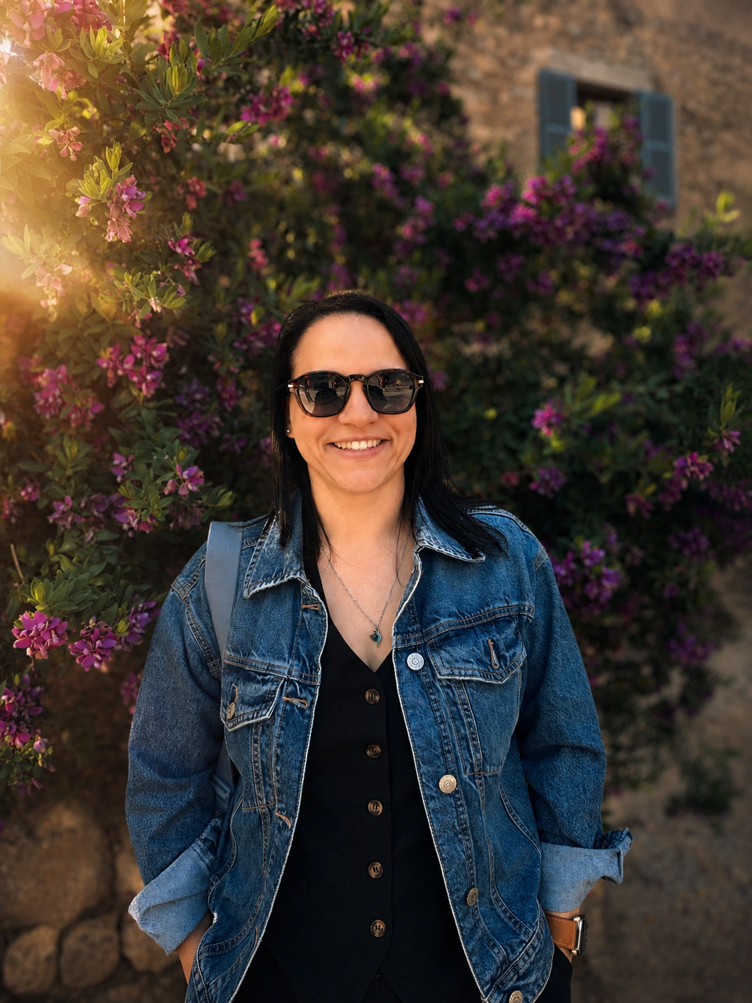 A woman smiling outdoors in front of flowering purple bushes, wearing sunglasses, a denim jacket, and a black shirt.