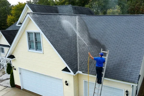 A person in a blue shirt and cap cleaning the roof of a yellow house with a power washer, standing on a ladder.