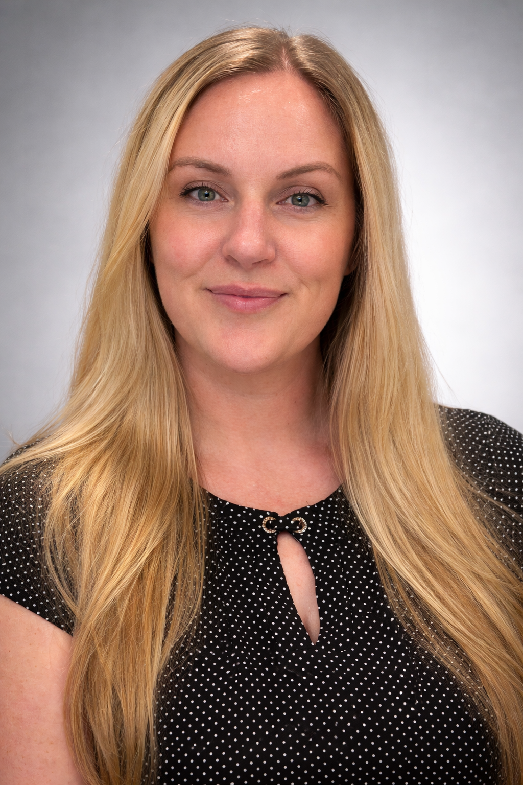 A woman with long blonde hair, blue eyes, and a slight smile, wearing a black polka dot blouse with a keyhole neckline, standing against a plain light gray background.