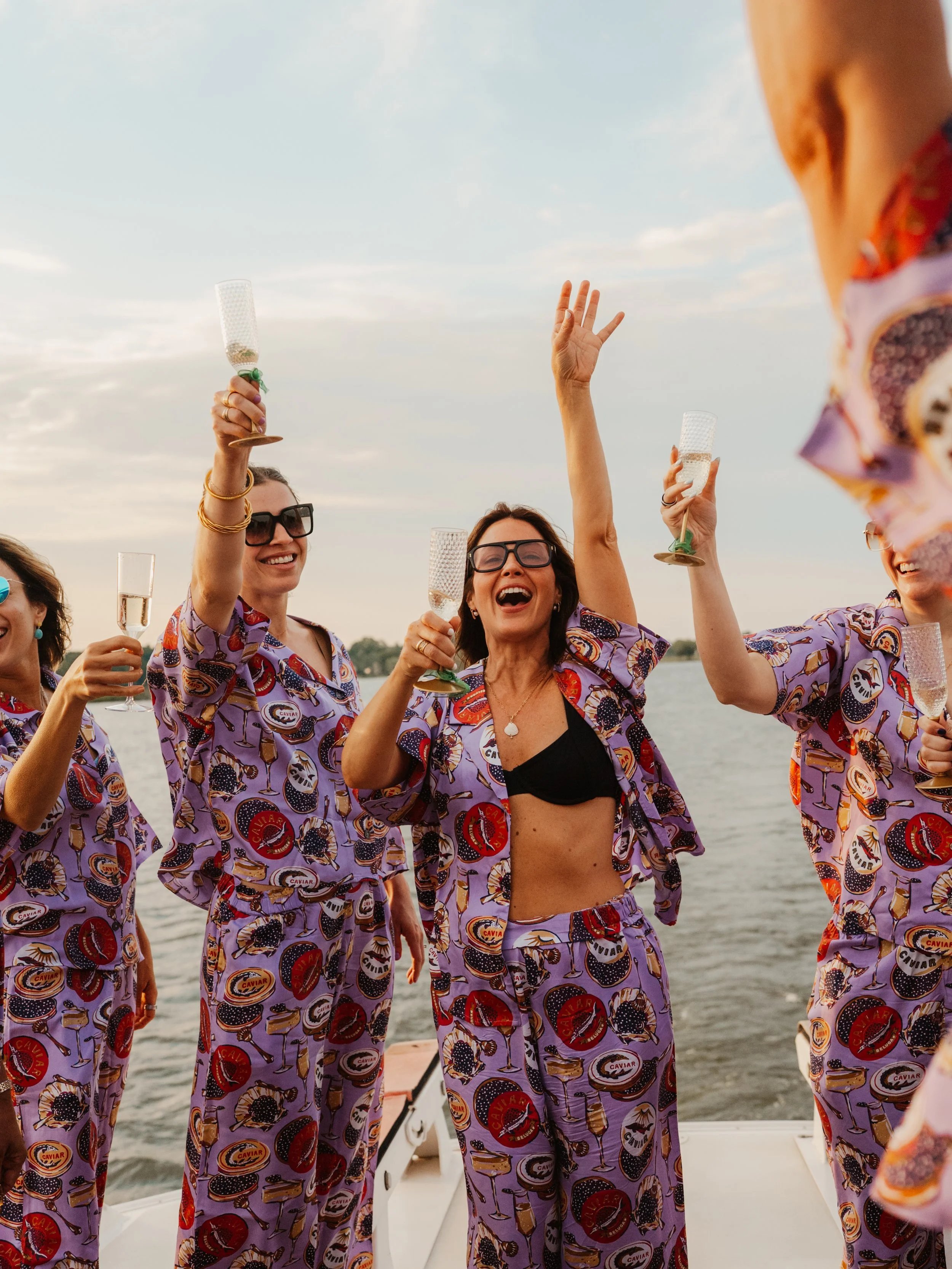 A group of people on a boat celebrating, wearing matching purple pajamas with caviar and champagne logos, holding glasses of champagne, smiling, with water and a cloudy sky in the background.