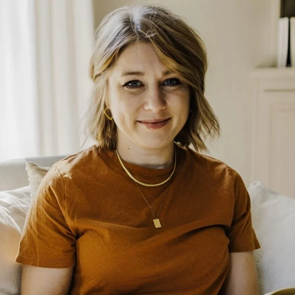 A woman with short, wavy blonde hair sitting on a beige couch, wearing a rust-colored shirt and gold jewelry, smiling at the camera in a cozy room with natural light.
