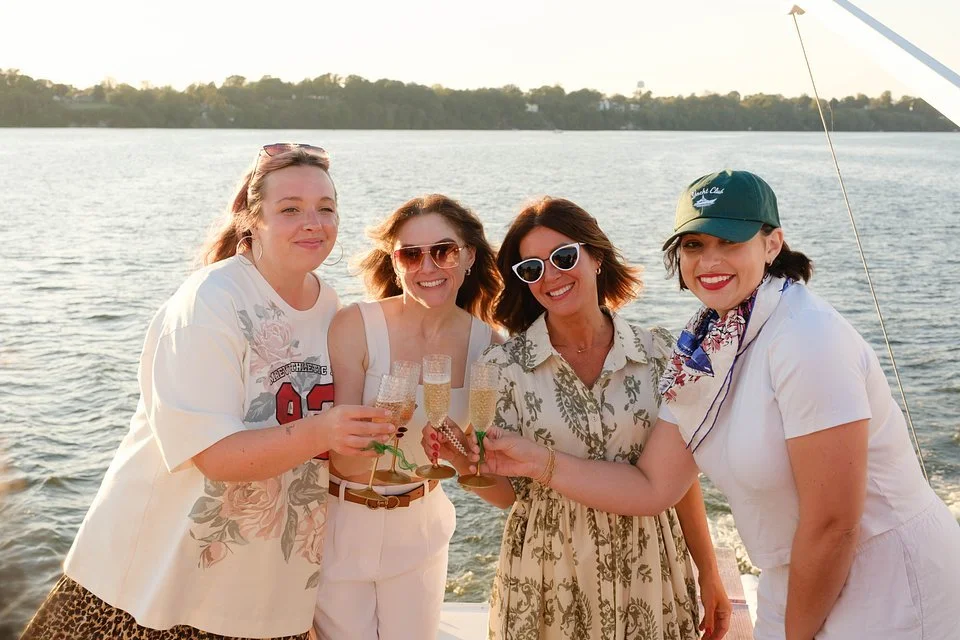 Four women on a boat holding glasses of champagne, smiling at the camera with a body of water and trees in the background.