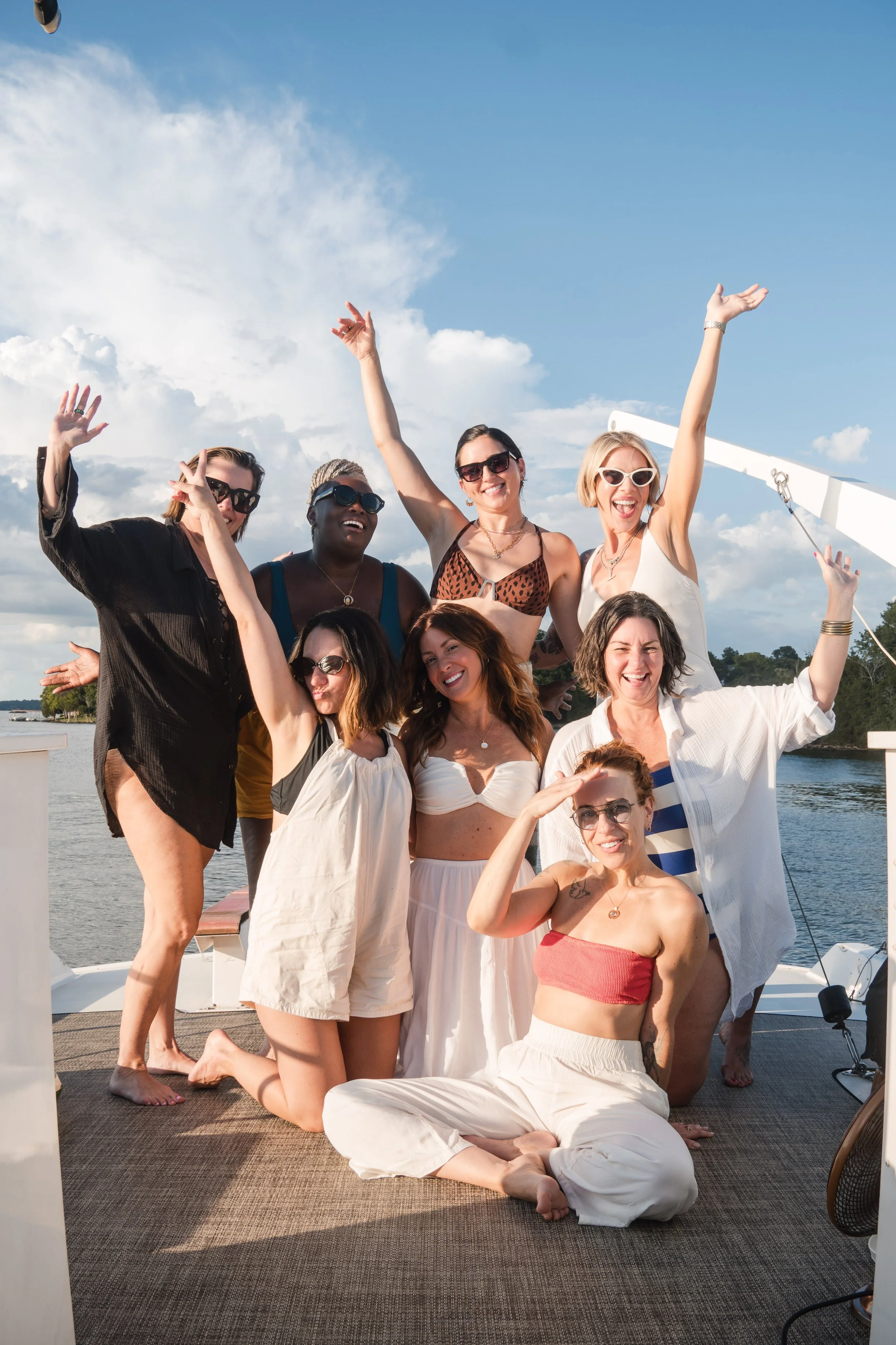 Group of women on a boat during the daytime, some are standing and others sitting or kneeling, enjoying the sunny weather with a river and blue sky with clouds in the background.