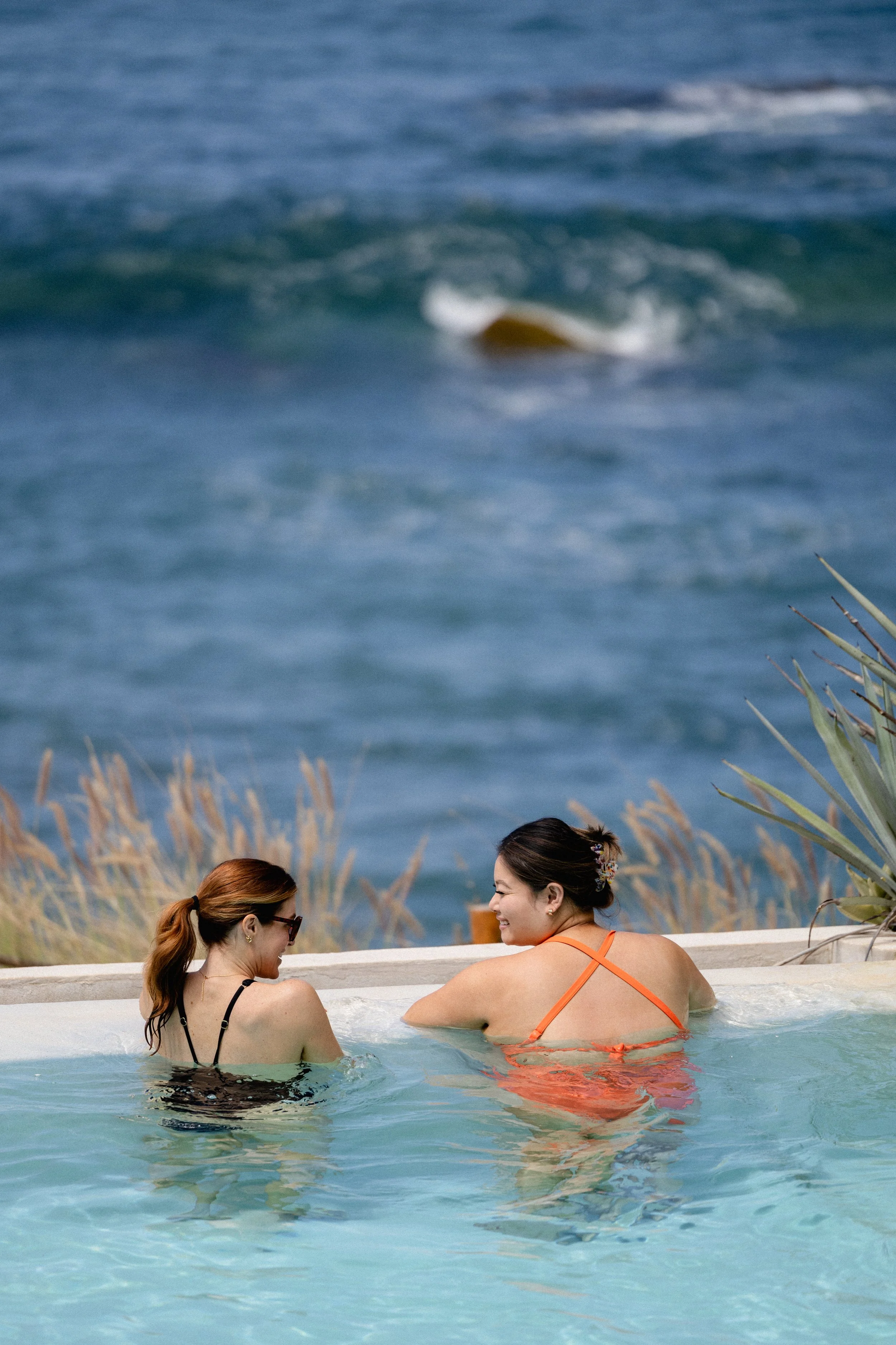 Two women sitting in a hot tub or pool, smiling and talking, with ocean waves in the background and some plants and grass nearby.