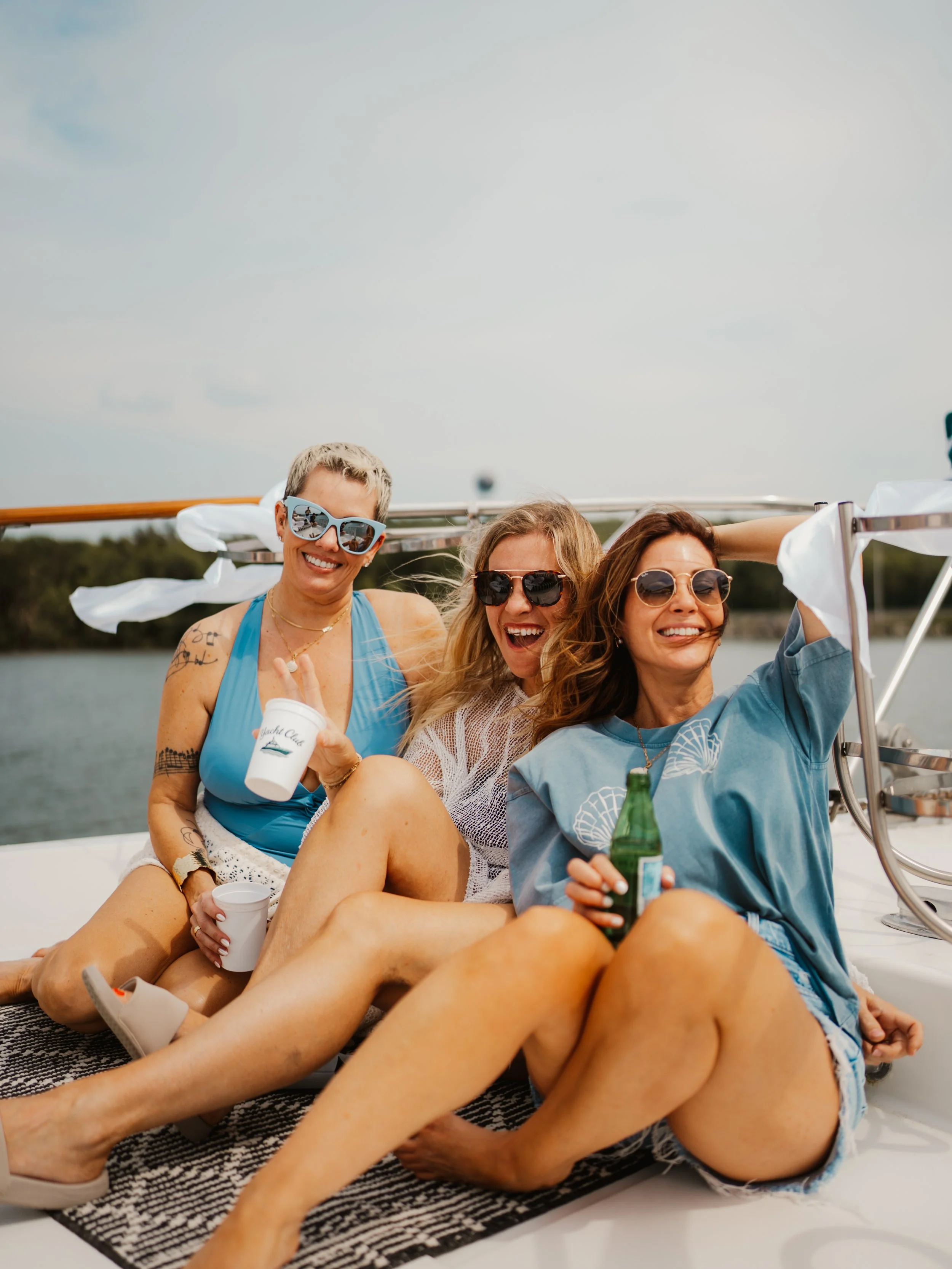 Four women sitting on a boat, smiling and enjoying drinks, with water and trees in the background.