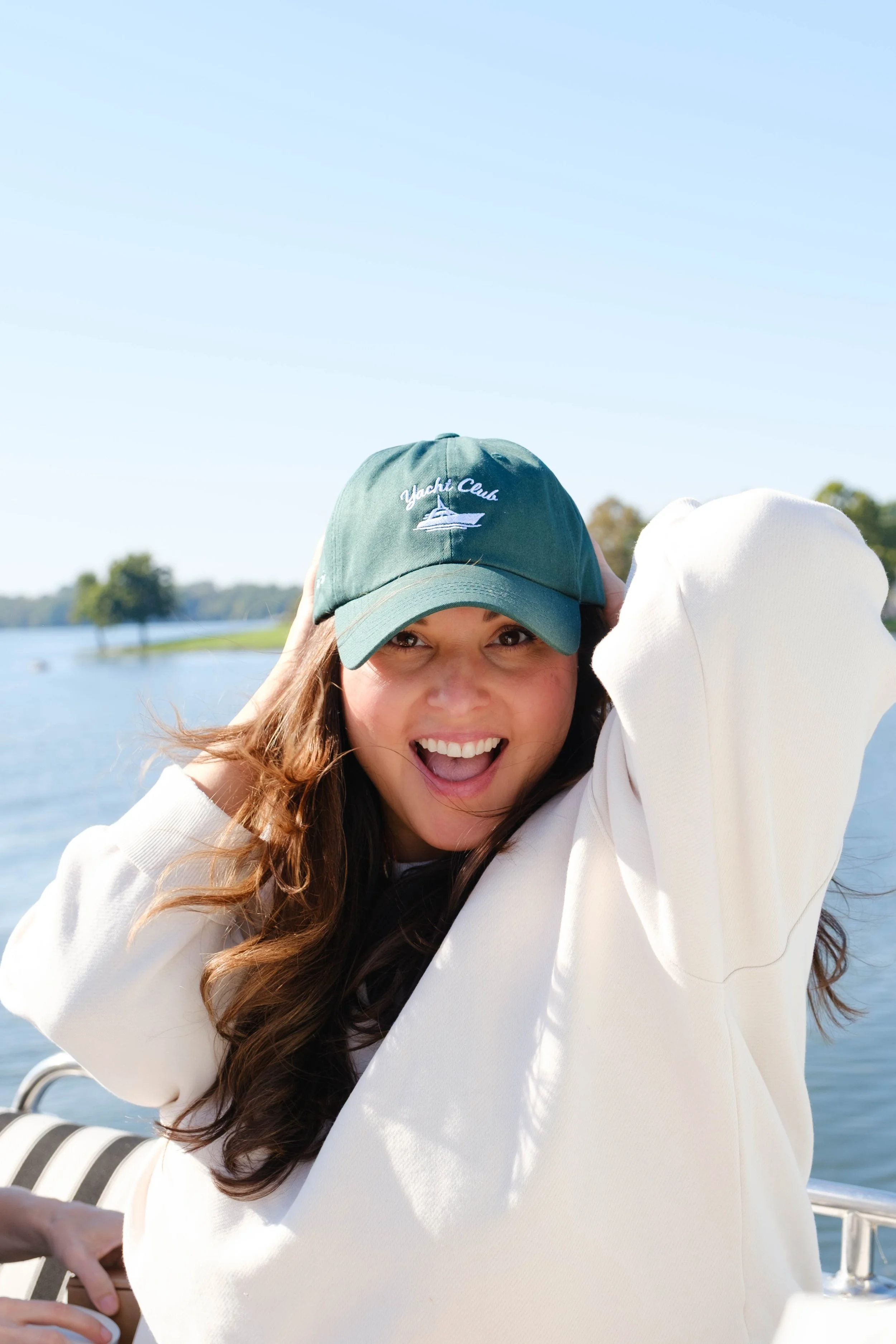 Close-up of a woman with long curly hair, wearing a green baseball cap with a boat and the words 'Yacht Club' embroidered on it, smiling and squinting in the sunlight near water, during daytime.