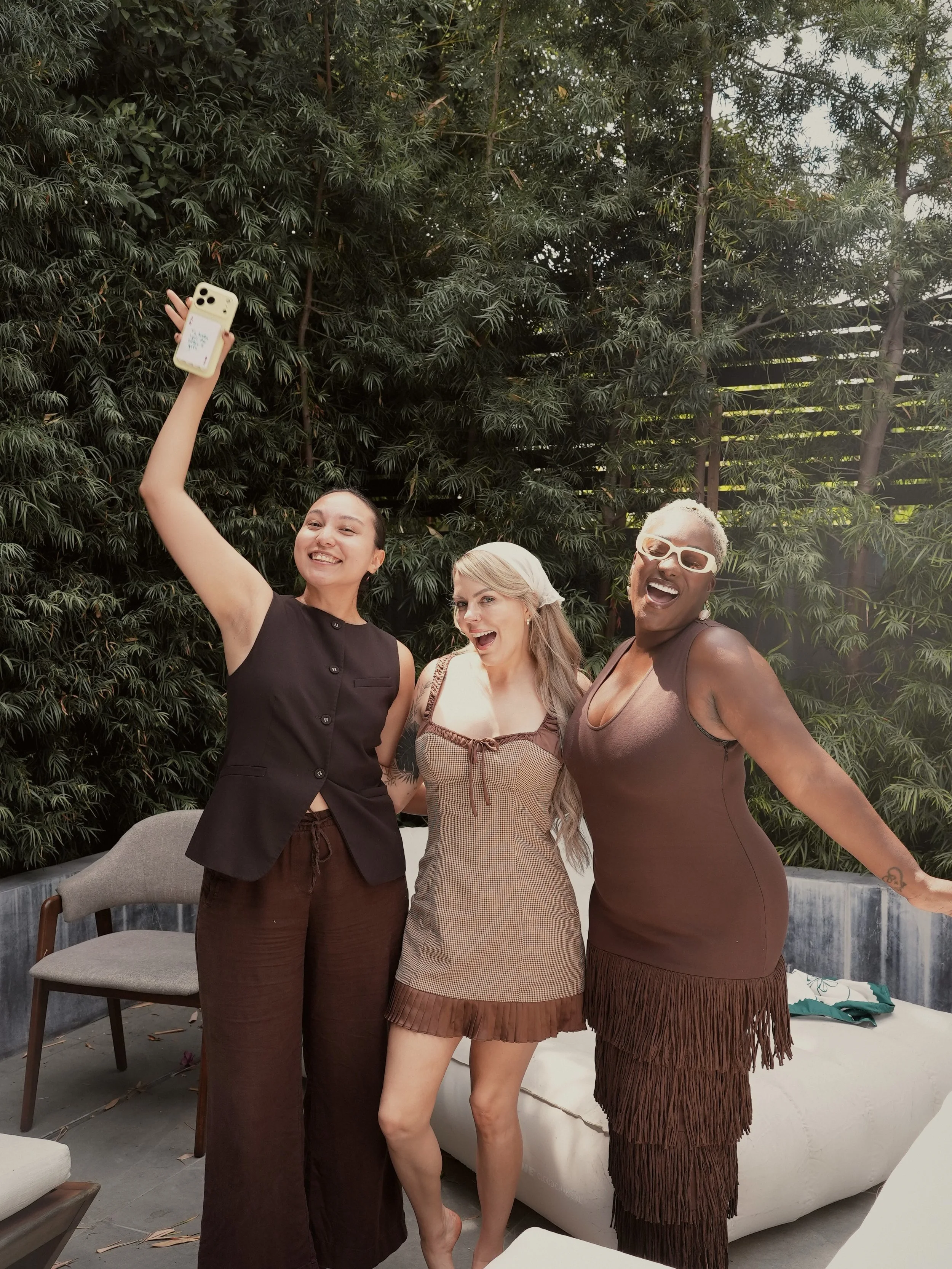 Three women and one person with short gray hair taking a selfie in front of a green leafy background. They are smiling and dressed in casual clothing.