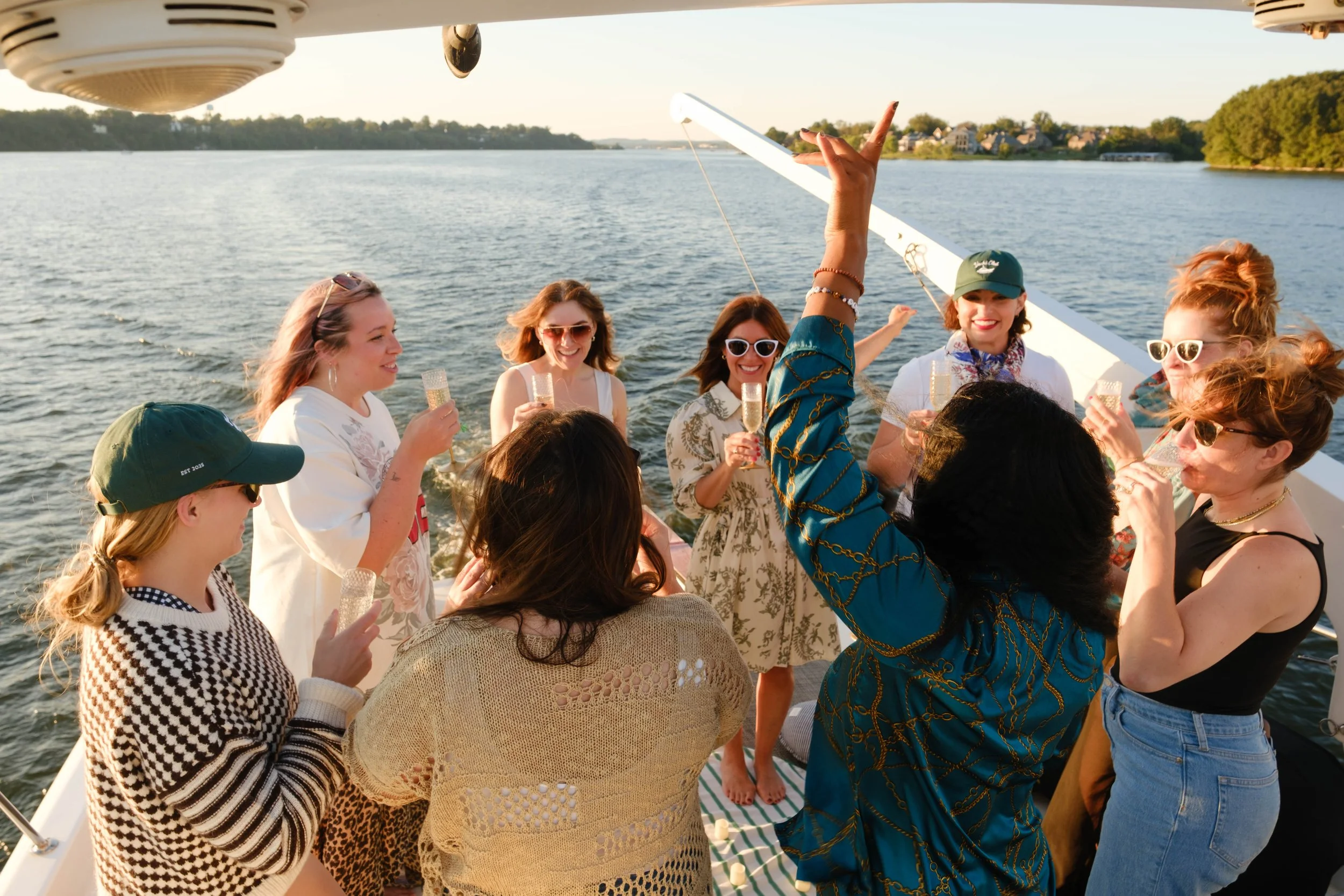 Group of women enjoying drinks on a boat with water and trees in the background during sunset.