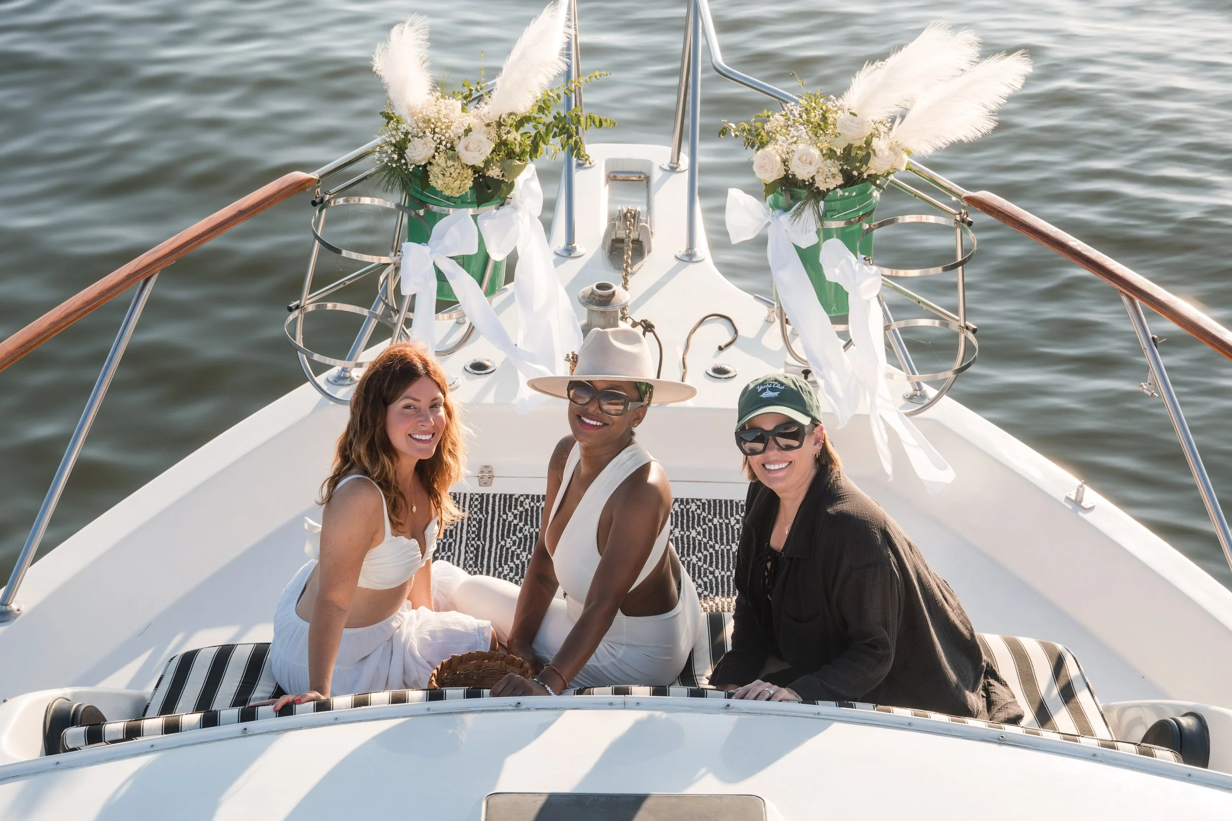 Four women sitting on the deck of a boat in the water, smiling and enjoying each other's company. The boat is decorated with white flowers and ribbons for a celebration.