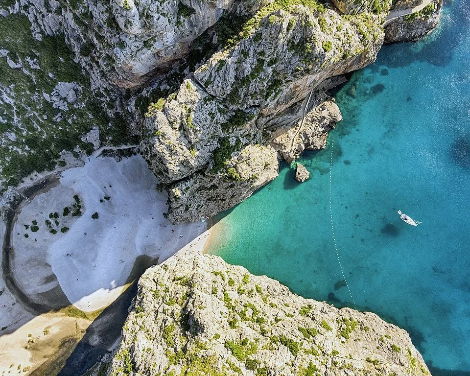 Aerial view of a cove with turquoise water, a boat floating, rocky cliffs, and a small sandy beach with a path leading to the cliffs.