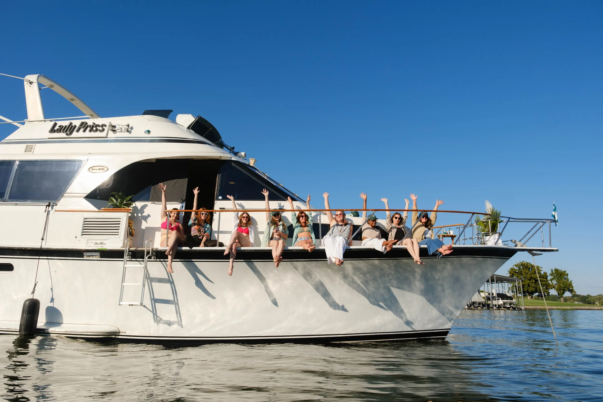 Group of people sitting on the deck of a large white yacht named Lady Priss, raising their hands, under a clear blue sky, with water and trees in the background.