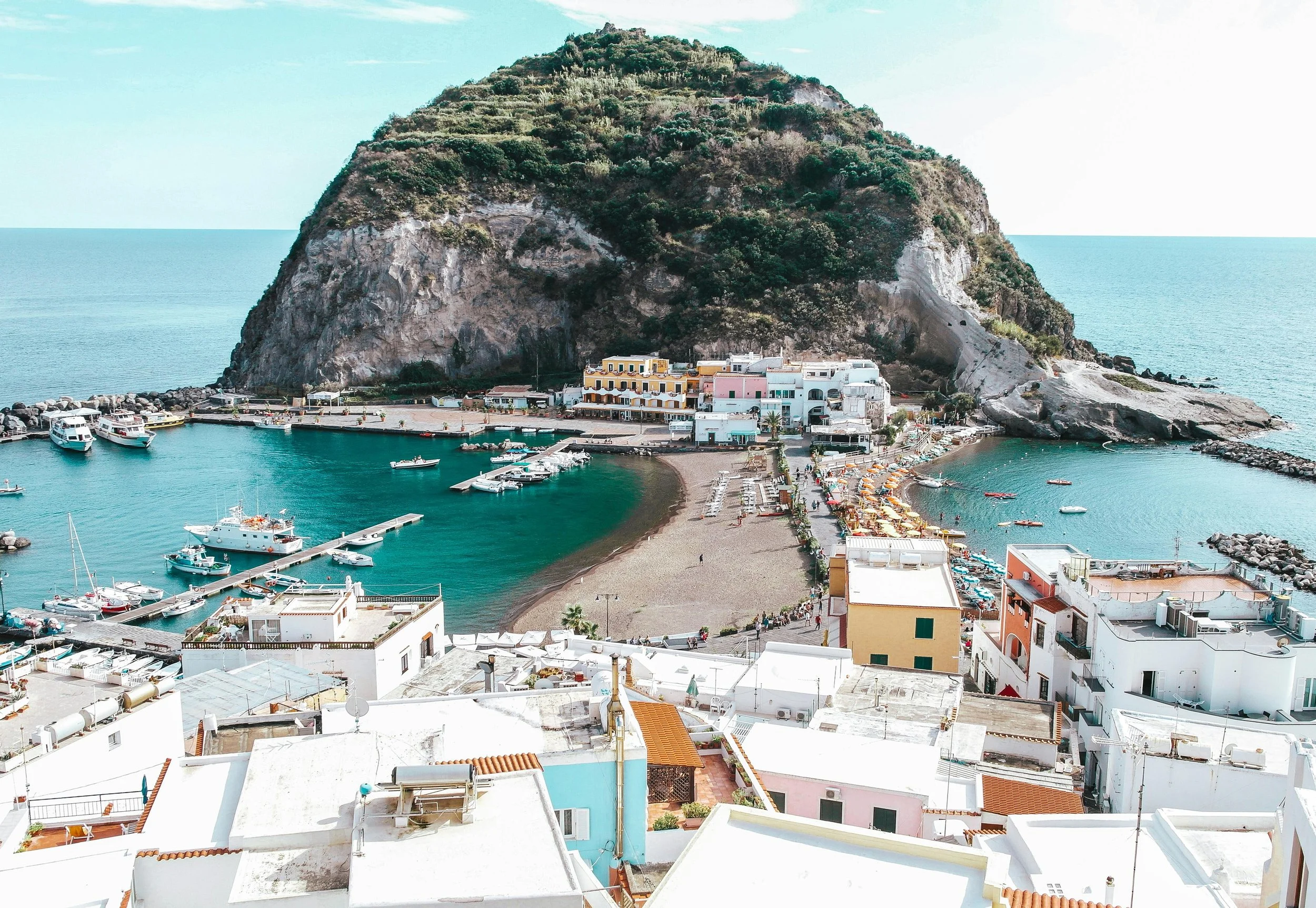 A coastal town with white buildings, a small harbor, and boats, set against a large rocky hill covered with greenery, overlooking the sea.