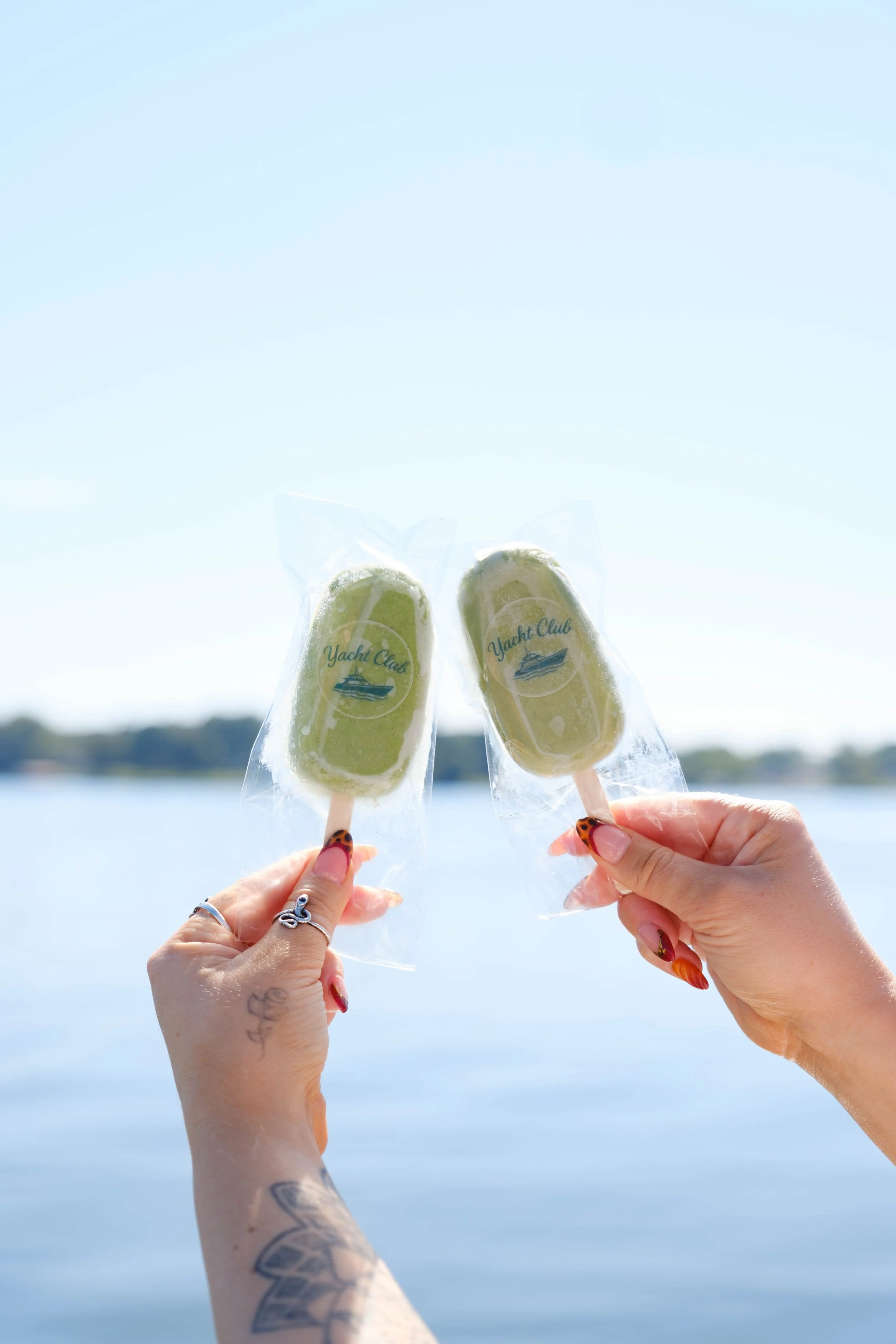 Two hands holding popsicles with 'Yacht Club' labels in front of a body of water and a clear sky.