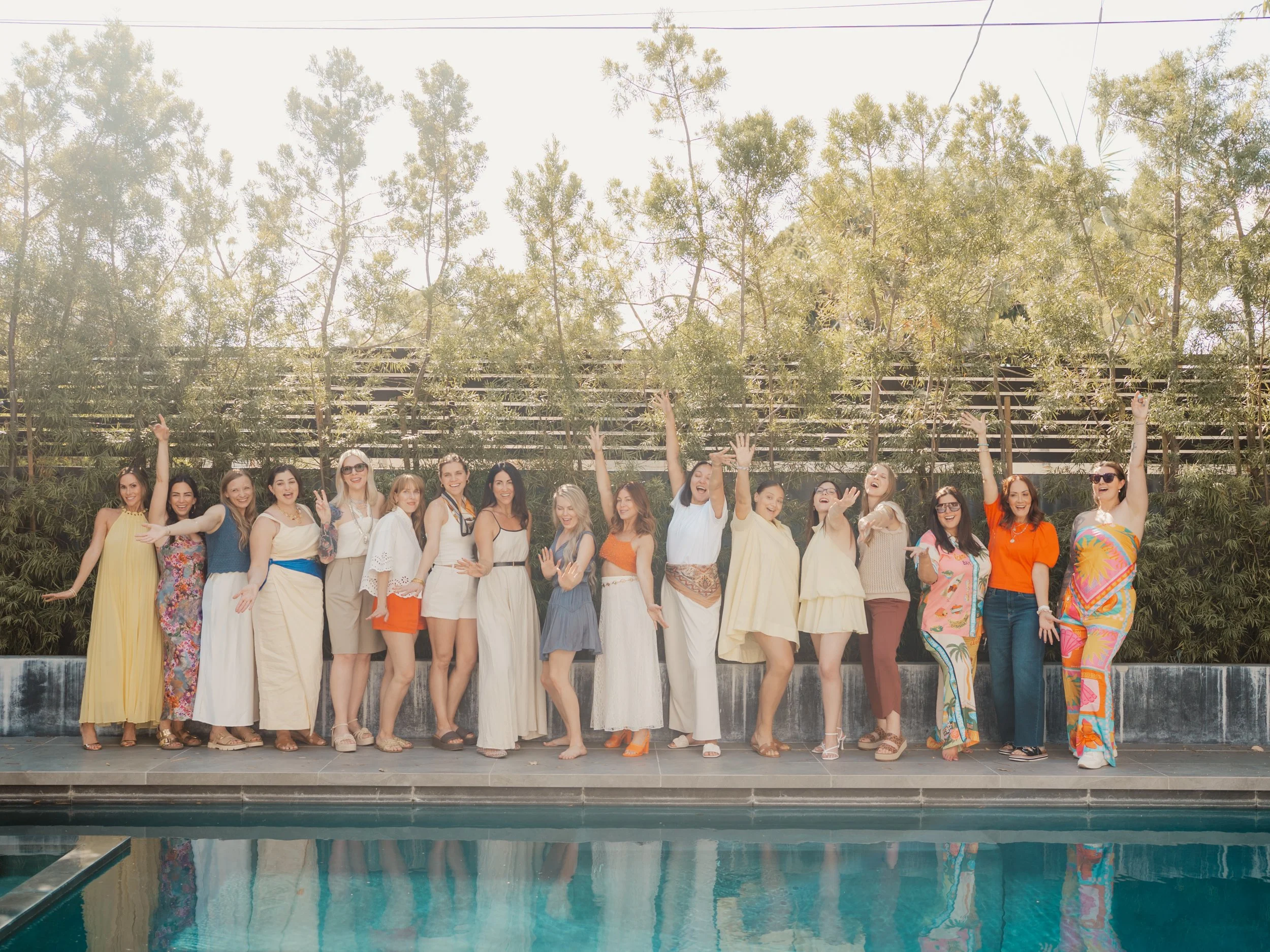 Group of diverse women standing on a ledge by a pool, smiling and raising their hands, outdoor setting with trees and a wooden fence in the background.