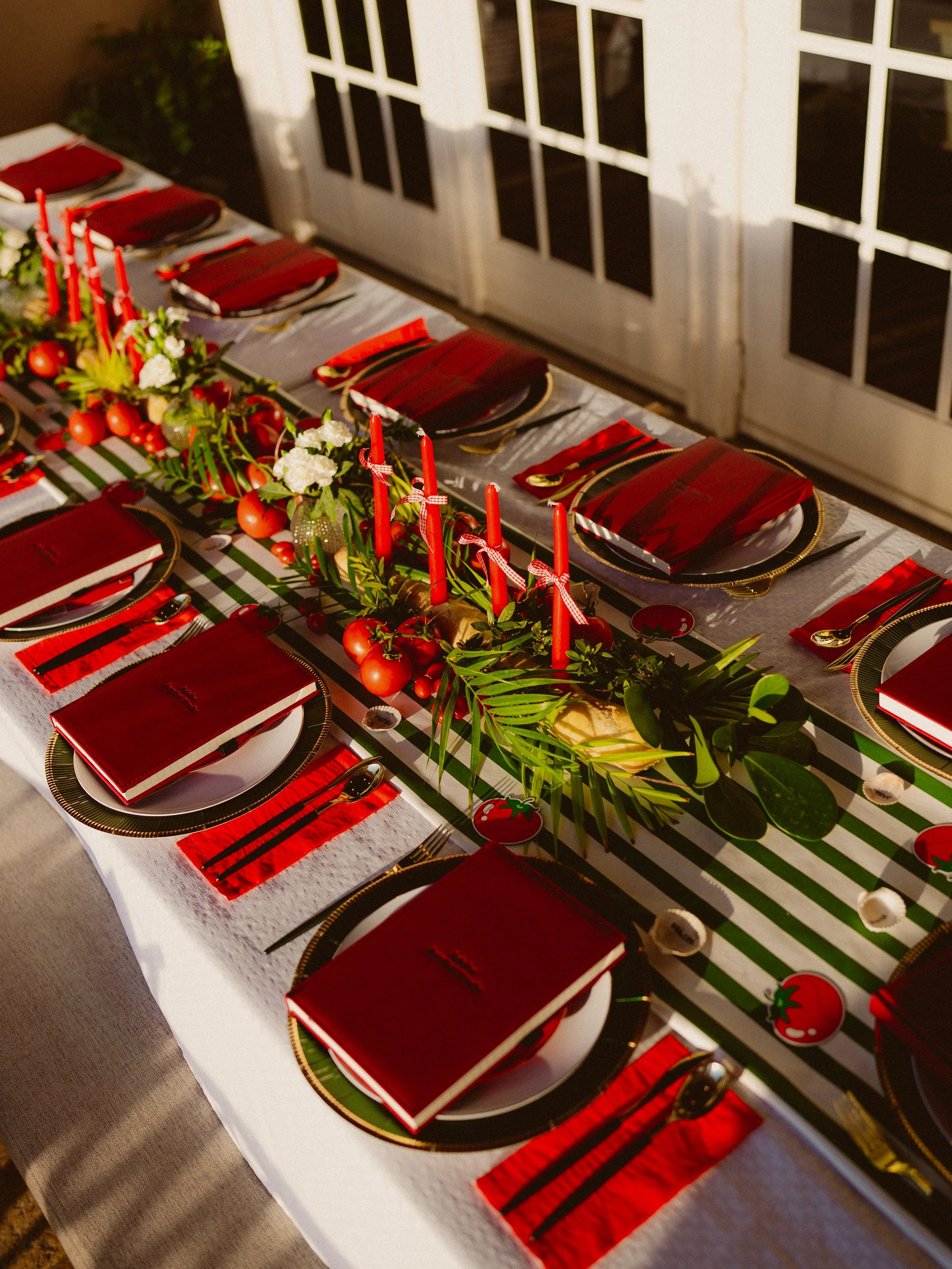 A festive table setting with red elements, black and gold plates, red napkins, and a centerpiece of greenery and red candles, likely prepared for a holiday celebration.