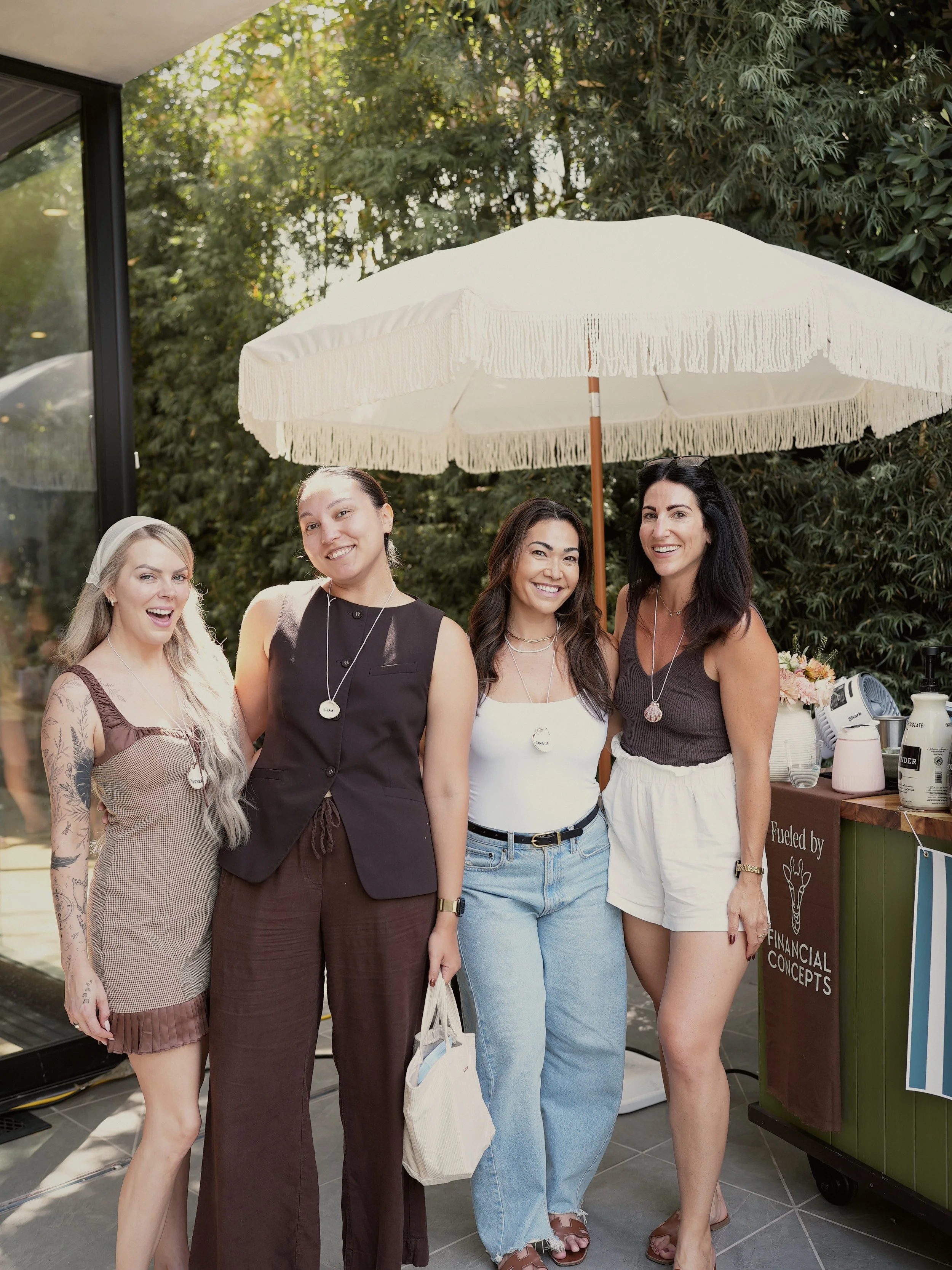 Four women standing outdoors under a large white umbrella, smiling for the photo. There is a green wooden stand with a sign and various items on it beside them, with trees in the background.