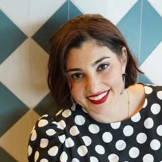 A woman with short brown hair, wearing a black and white polka dot top, smiling in front of a geometric patterned wall.