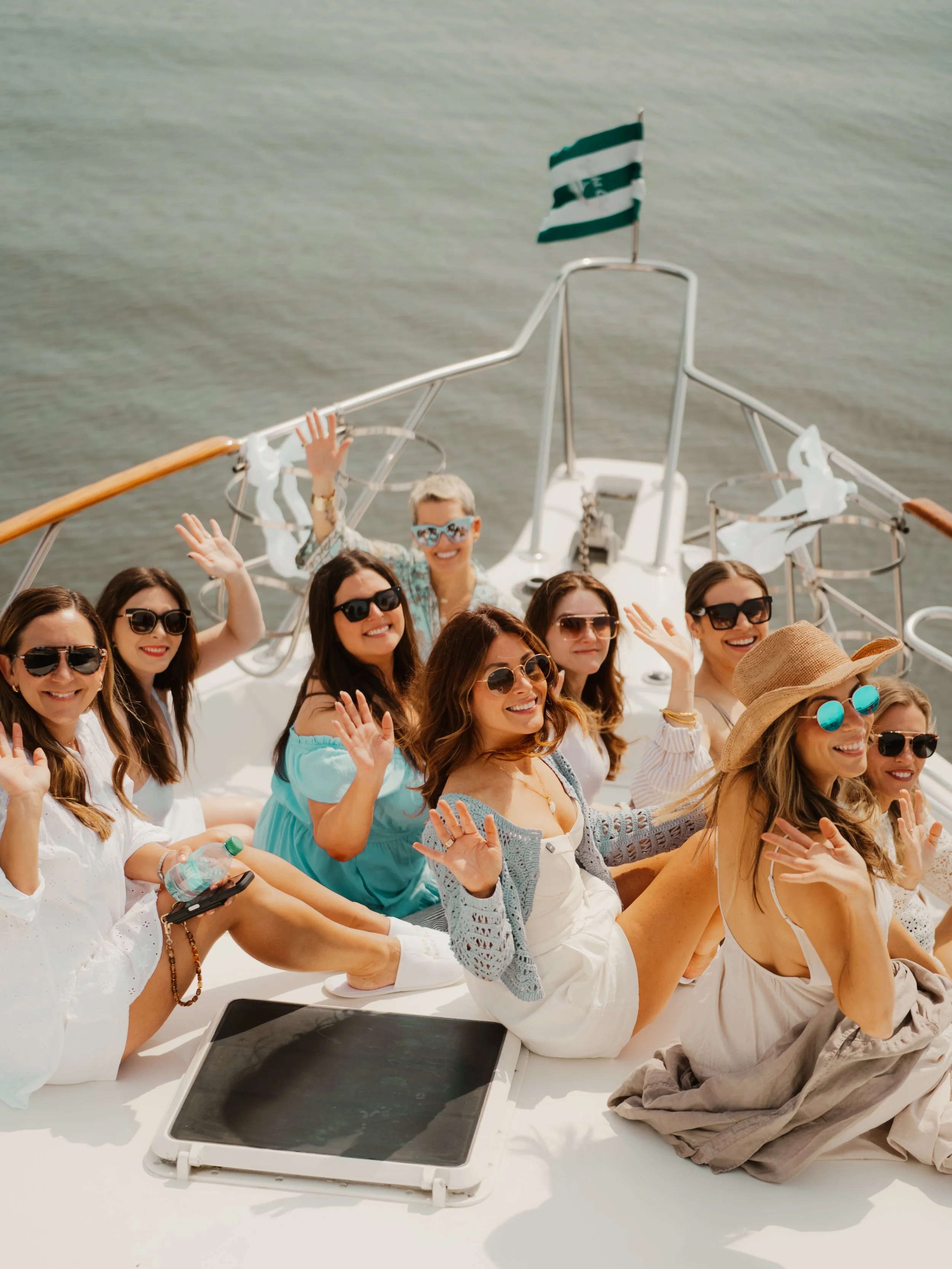 A group of smiling women wearing sunglasses and summer clothes, sitting on a boat, waving at the camera.