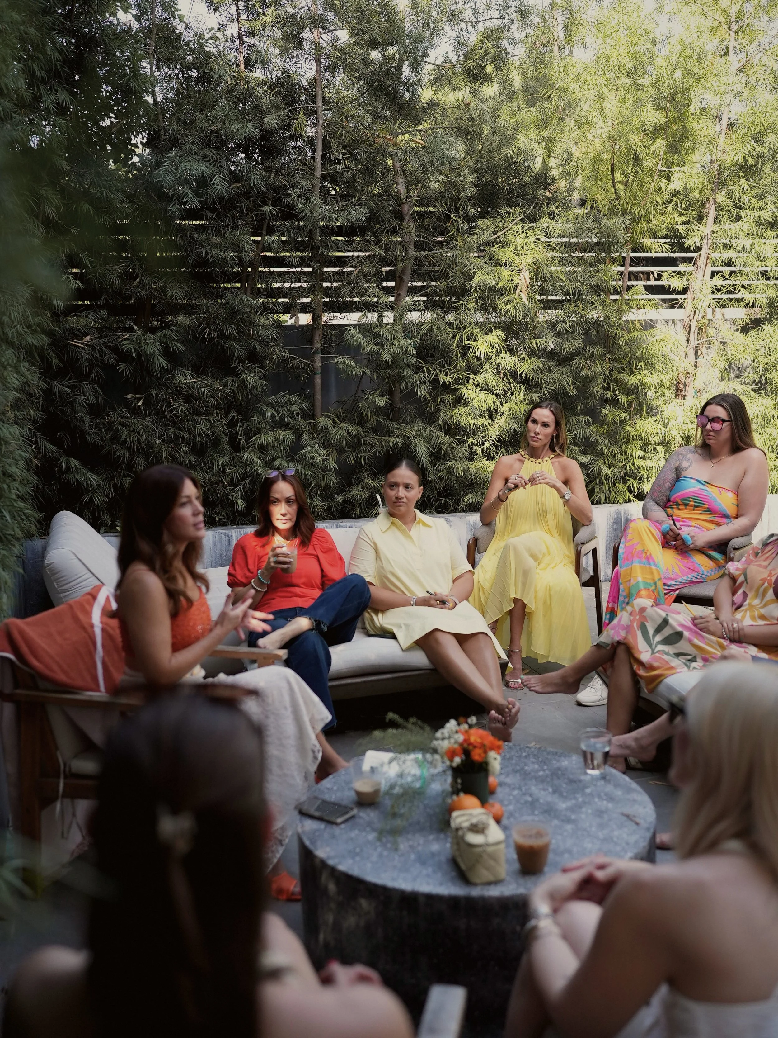 Group of women sitting outdoors on a patio, engaged in conversation, with flowers and drinks on a table in the center, and surrounded by green trees.