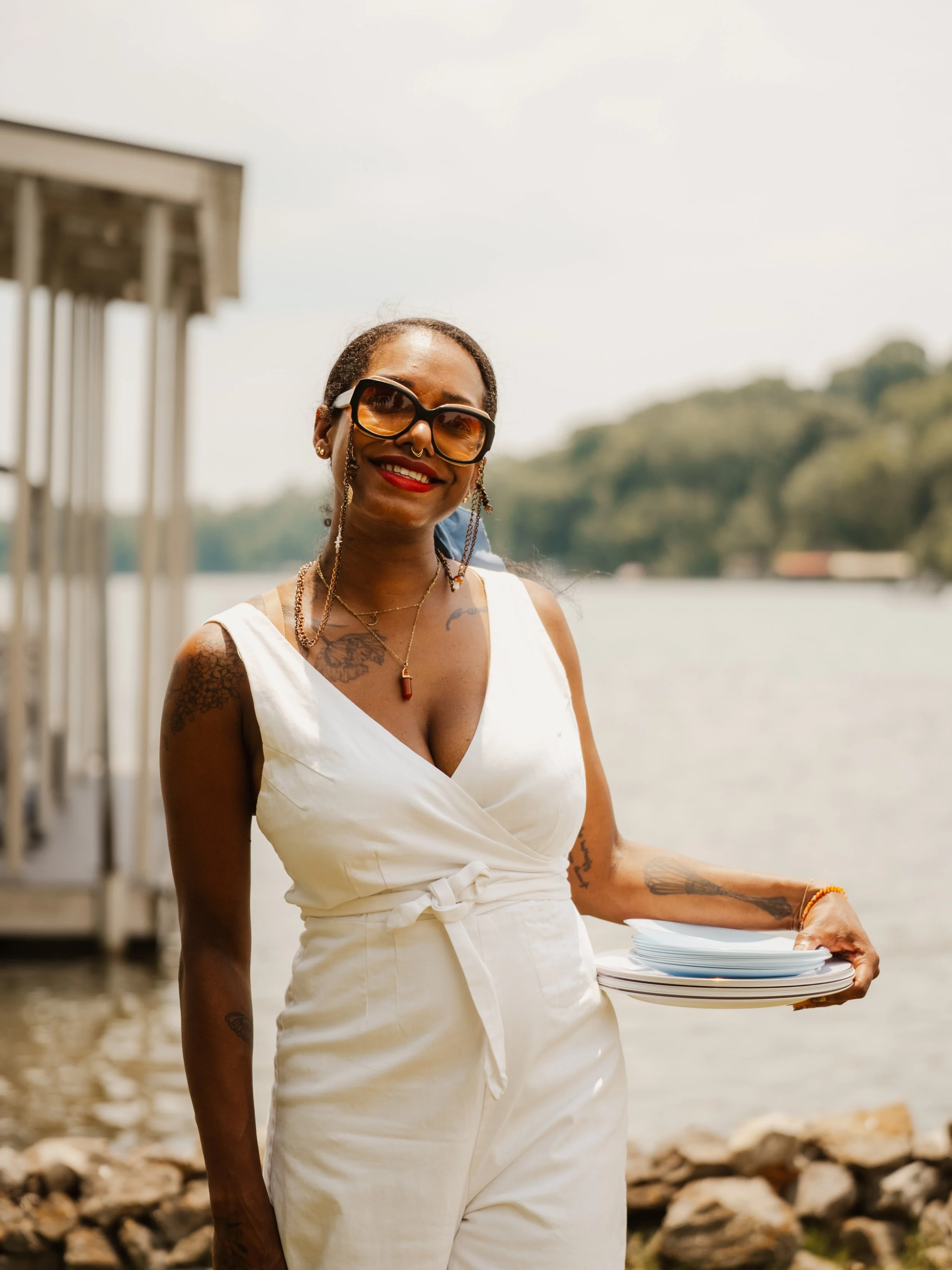 A smiling woman wearing sunglasses and a white dress holds a stack of plates outdoors near a body of water, with trees in the background.