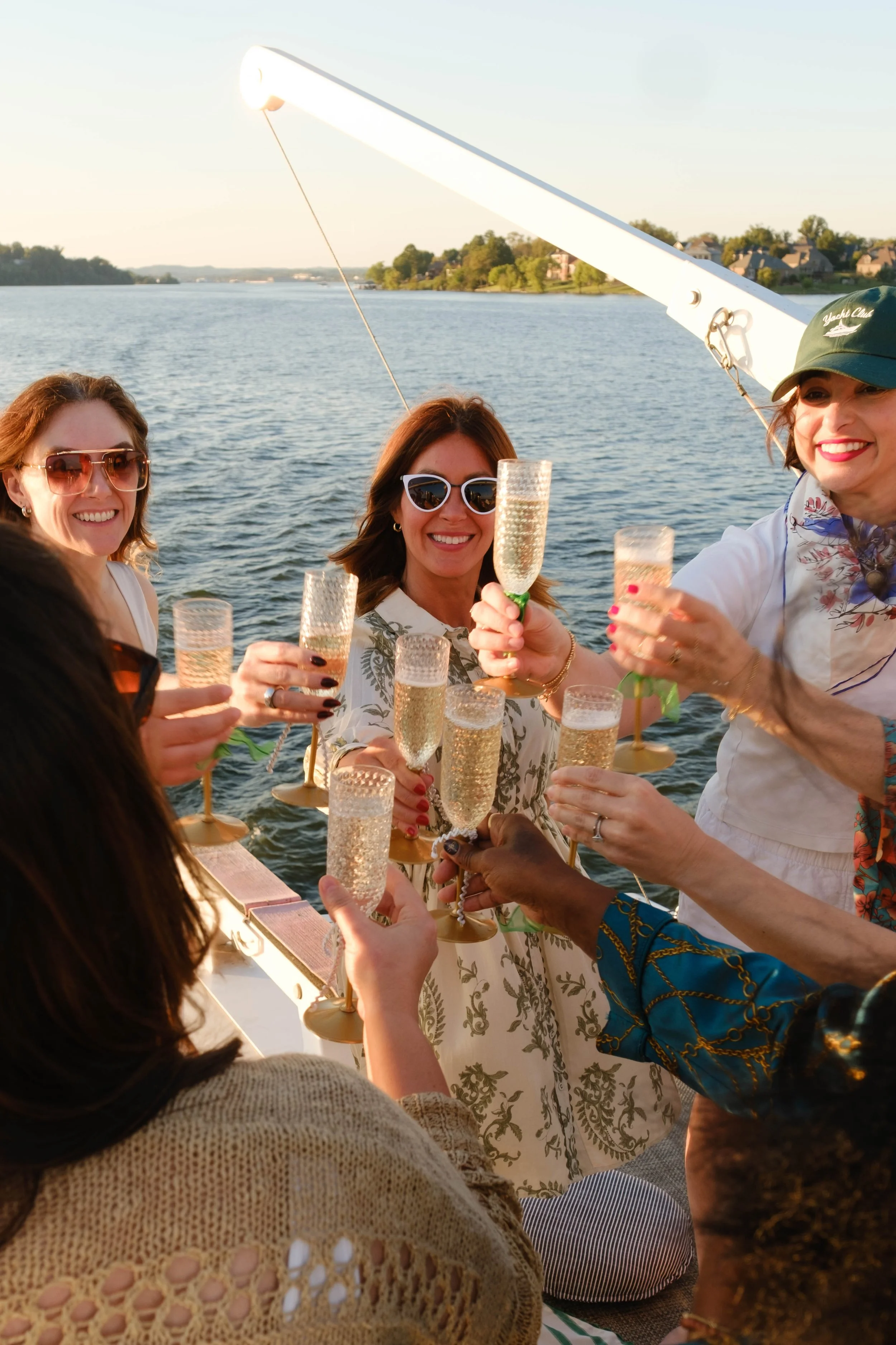 A group of women on a boat celebrating with champagne, smiling and holding glasses, with a river and houses in the background during sunset.