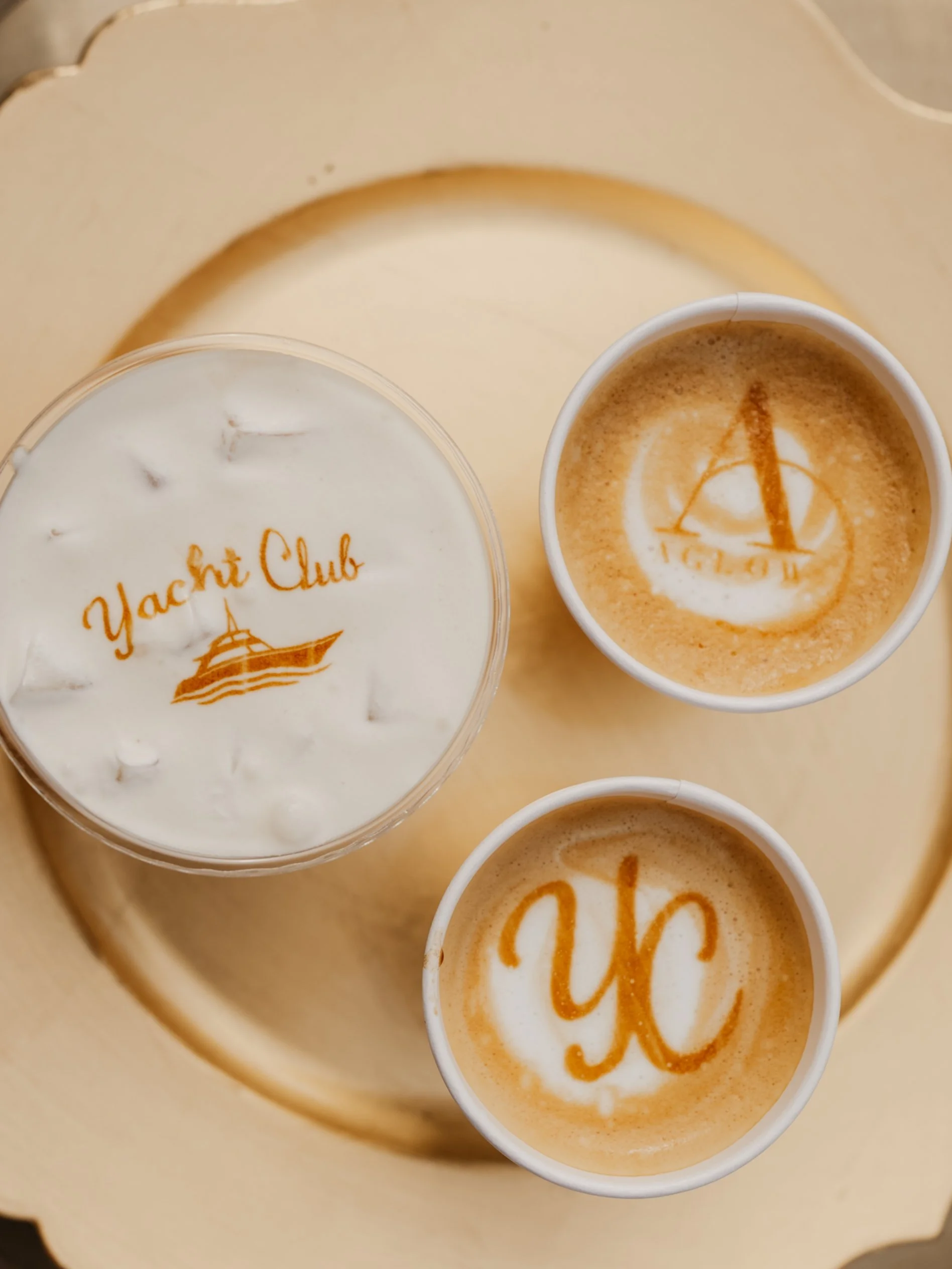 Close-up of three cups of coffee with latte art and the words 'Yacht Club' written on the foam, placed on a beige tray.