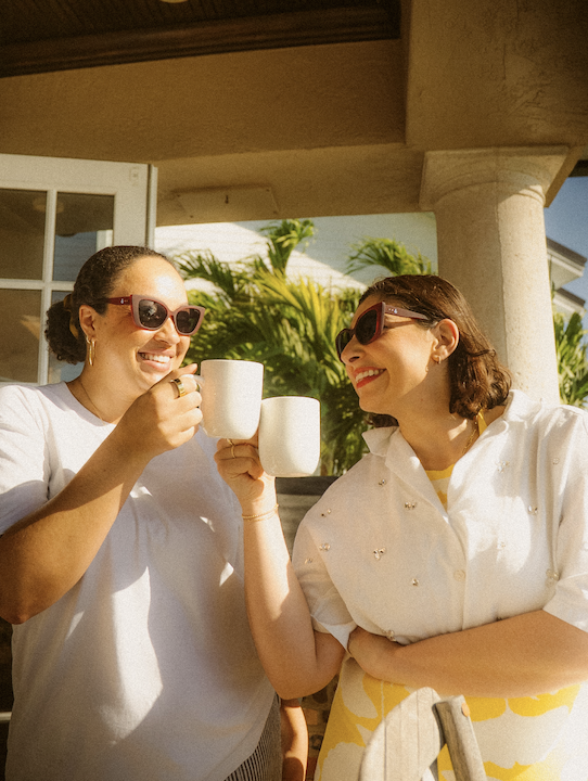 Two women with sunglasses smiling and clinking white mugs outdoors with a porch, green plants, and a house in the background.