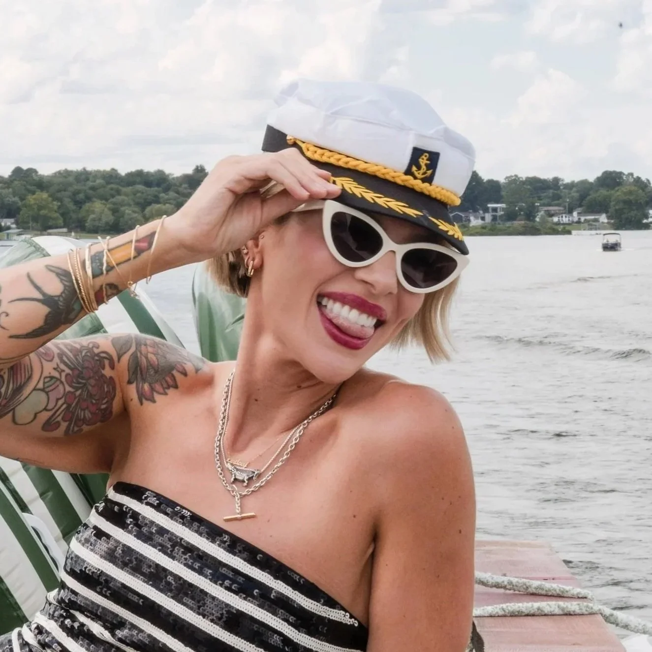 Smiling woman wearing sunglasses and a white captain's hat with a gold wreath, near a body of water with boats in the background.