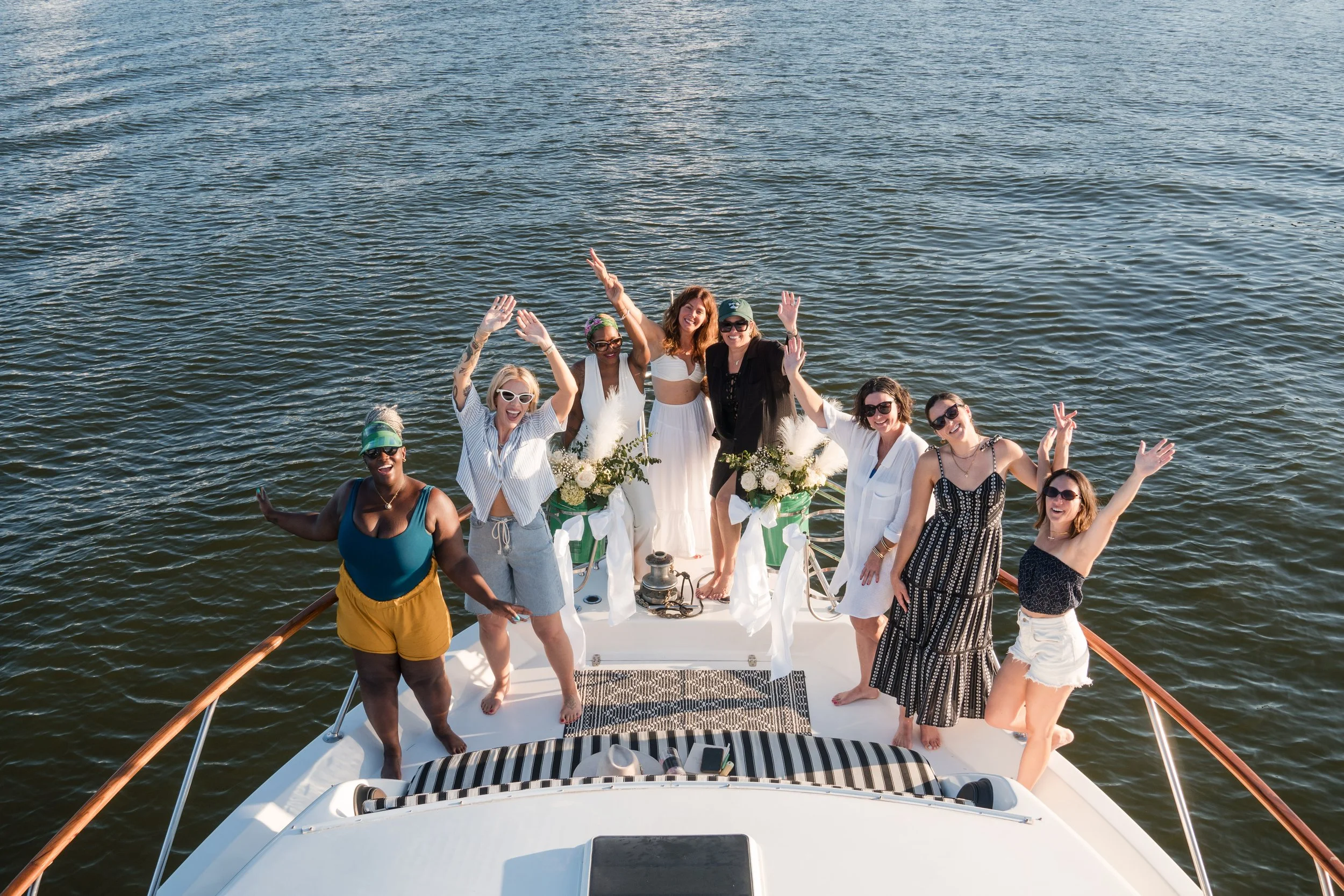 Group of women celebrating on a boat with water in the background; some women are wearing sunglasses and summer dresses, holding flowers and waving.