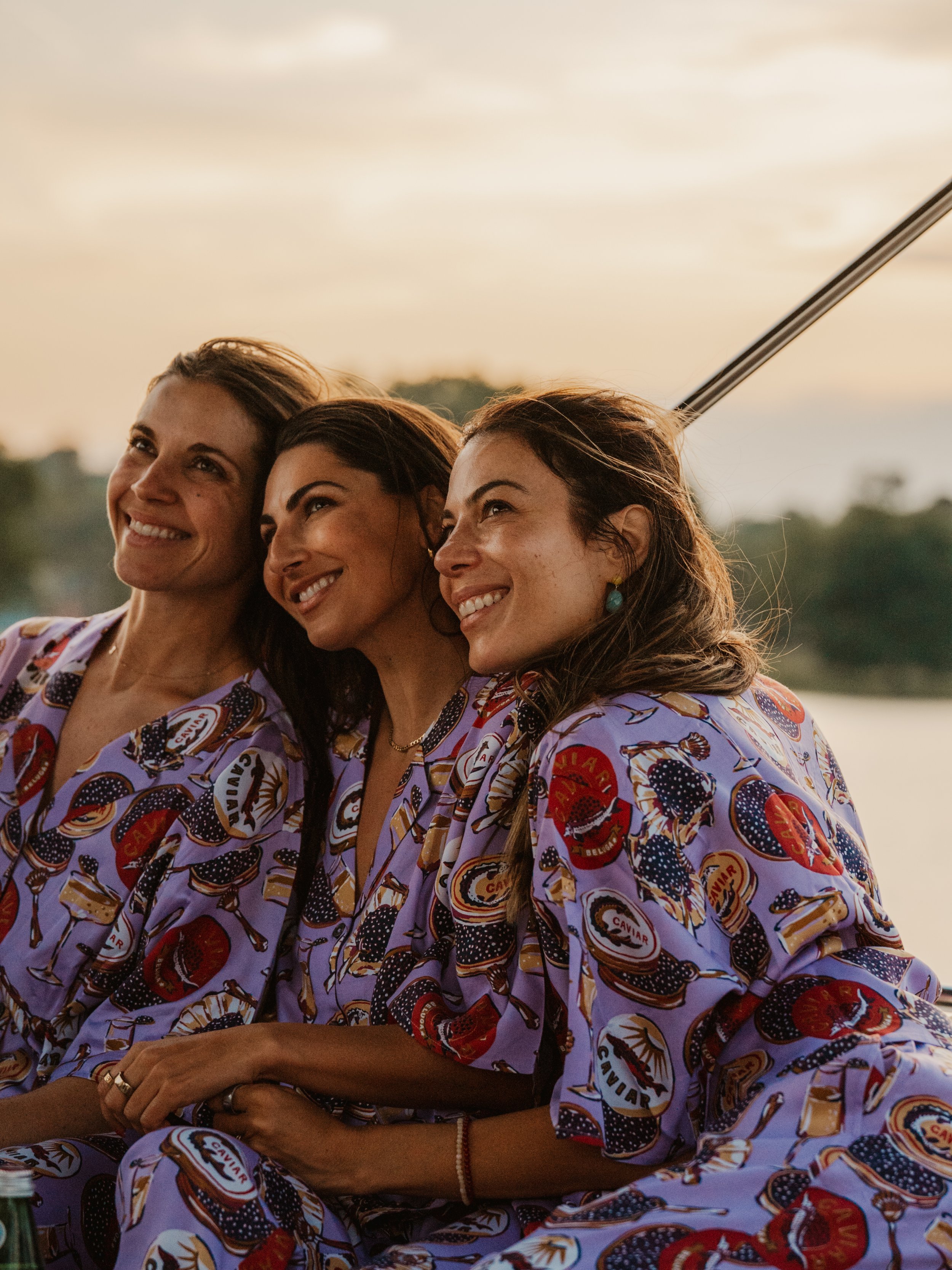Three women sitting outdoors during sunset, smiling and looking to the left, wearing matching patterned shirts with caviar and wine logos.