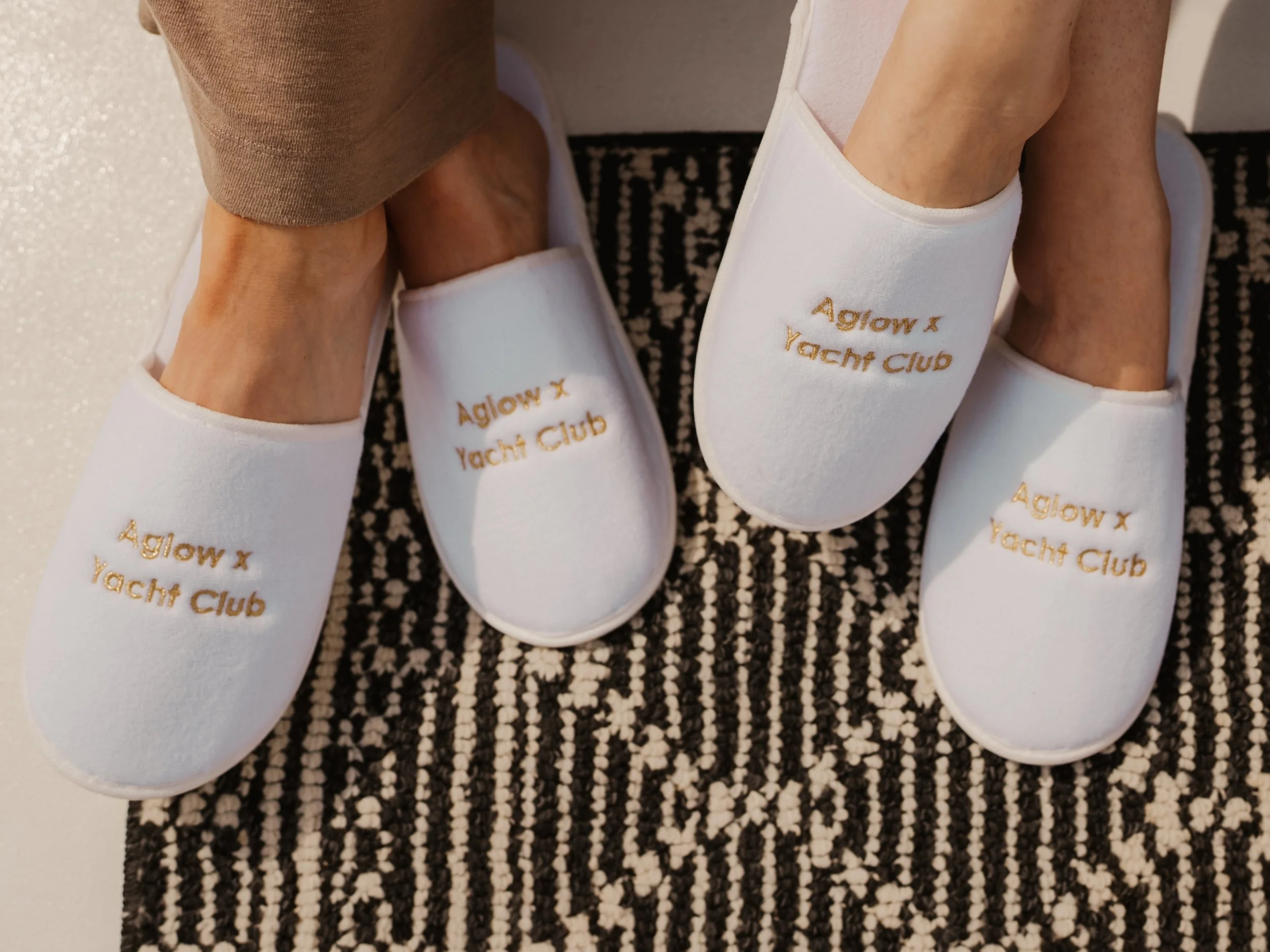 A pair of white slippers with gold embroidered text reading 'Aglow Yacht Club', placed on a black andwhite patterned rug, next to a person's legs and brown clothing.