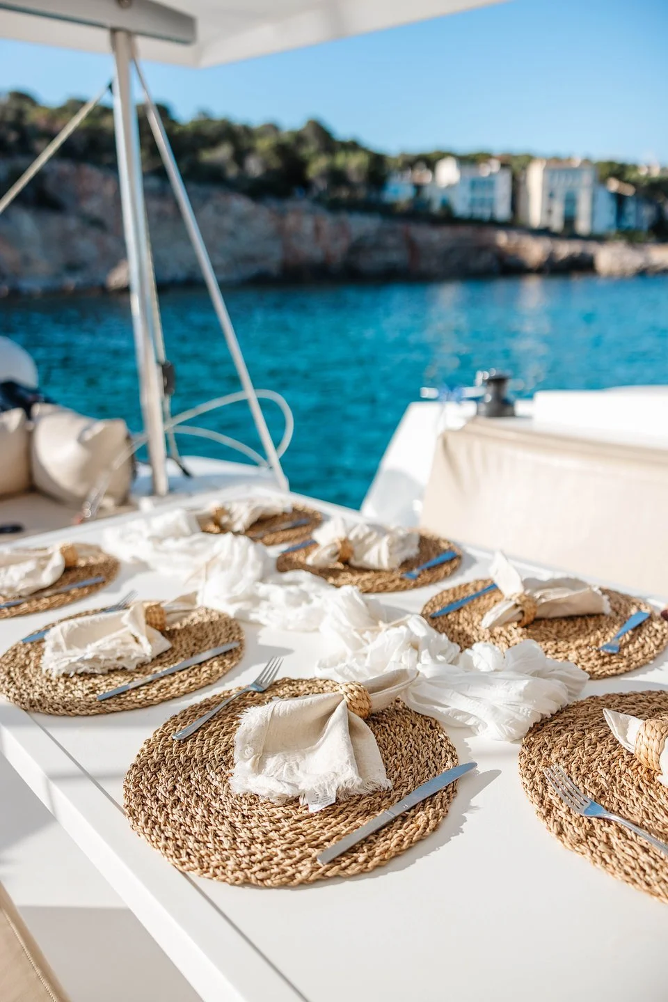 A table set for dining on a boat overlooking the water and distant buildings, with woven placemats, cutlery, and torn white napkins.