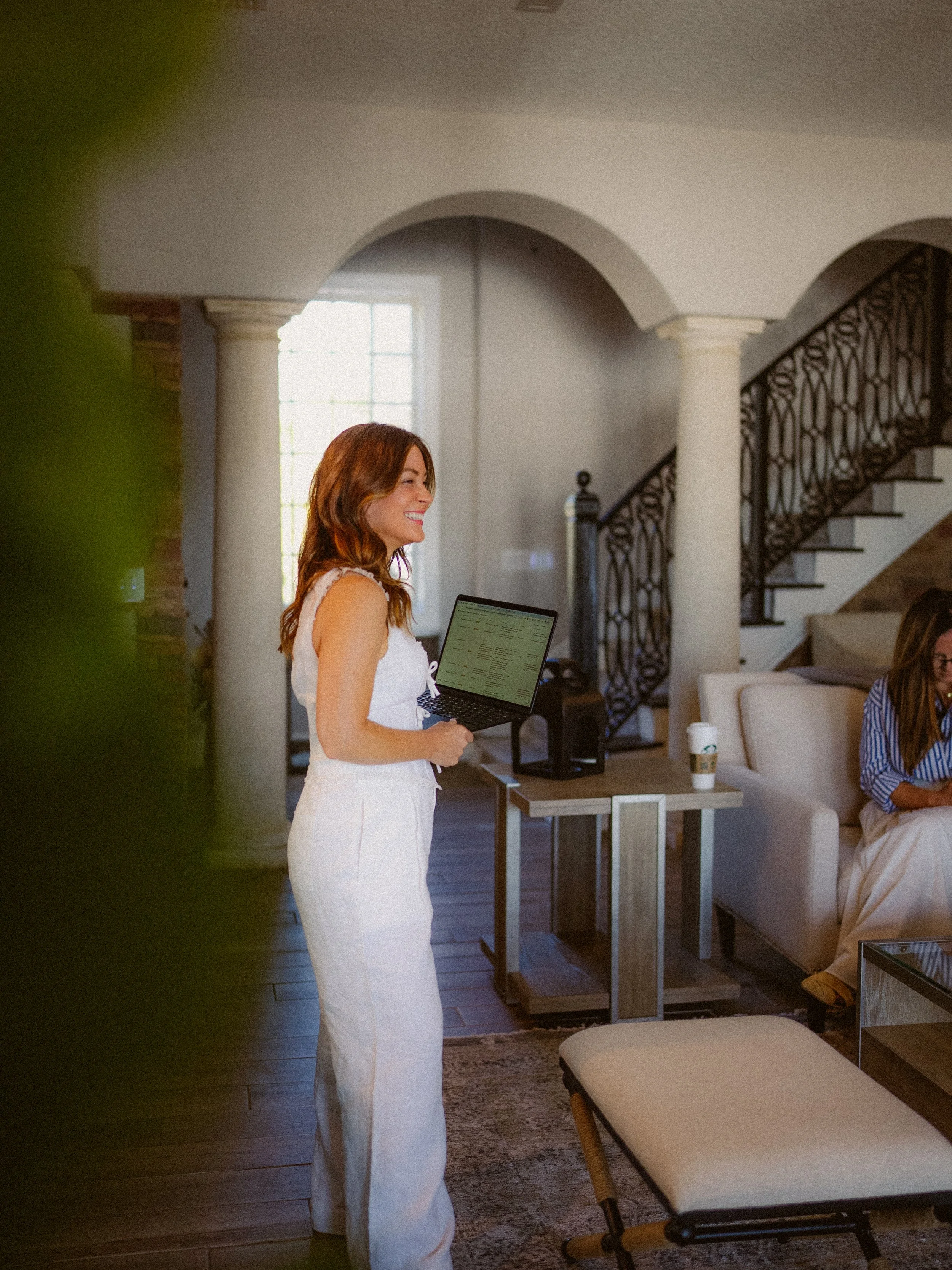 A woman with wavy red hair, wearing a white sleeveless top and white pants, smiling while holding a laptop inside a cozy living room with arched doorways and a staircase in the background.