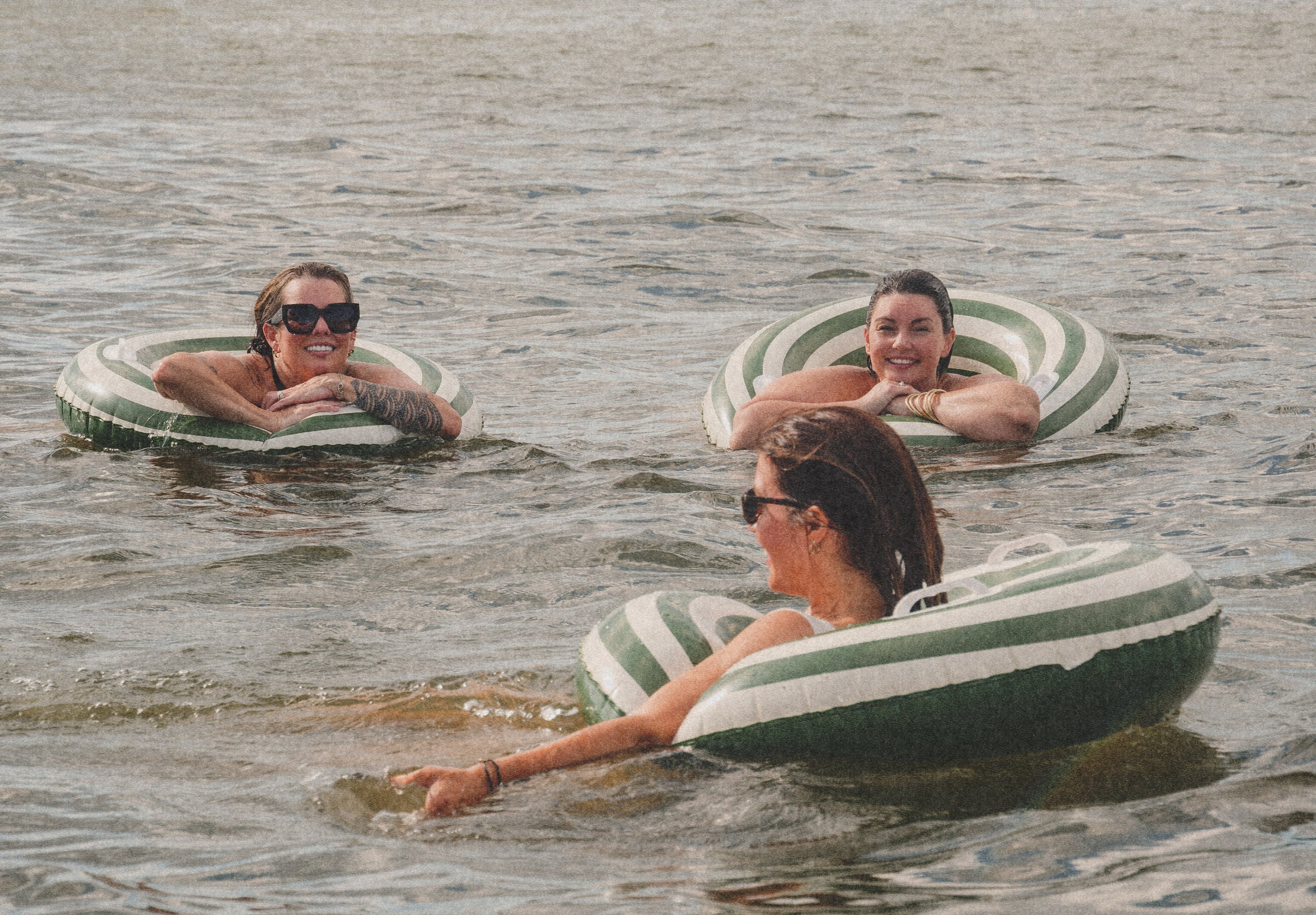 Two women lounging on green and white striped inflatable tubes in the water at the beach.