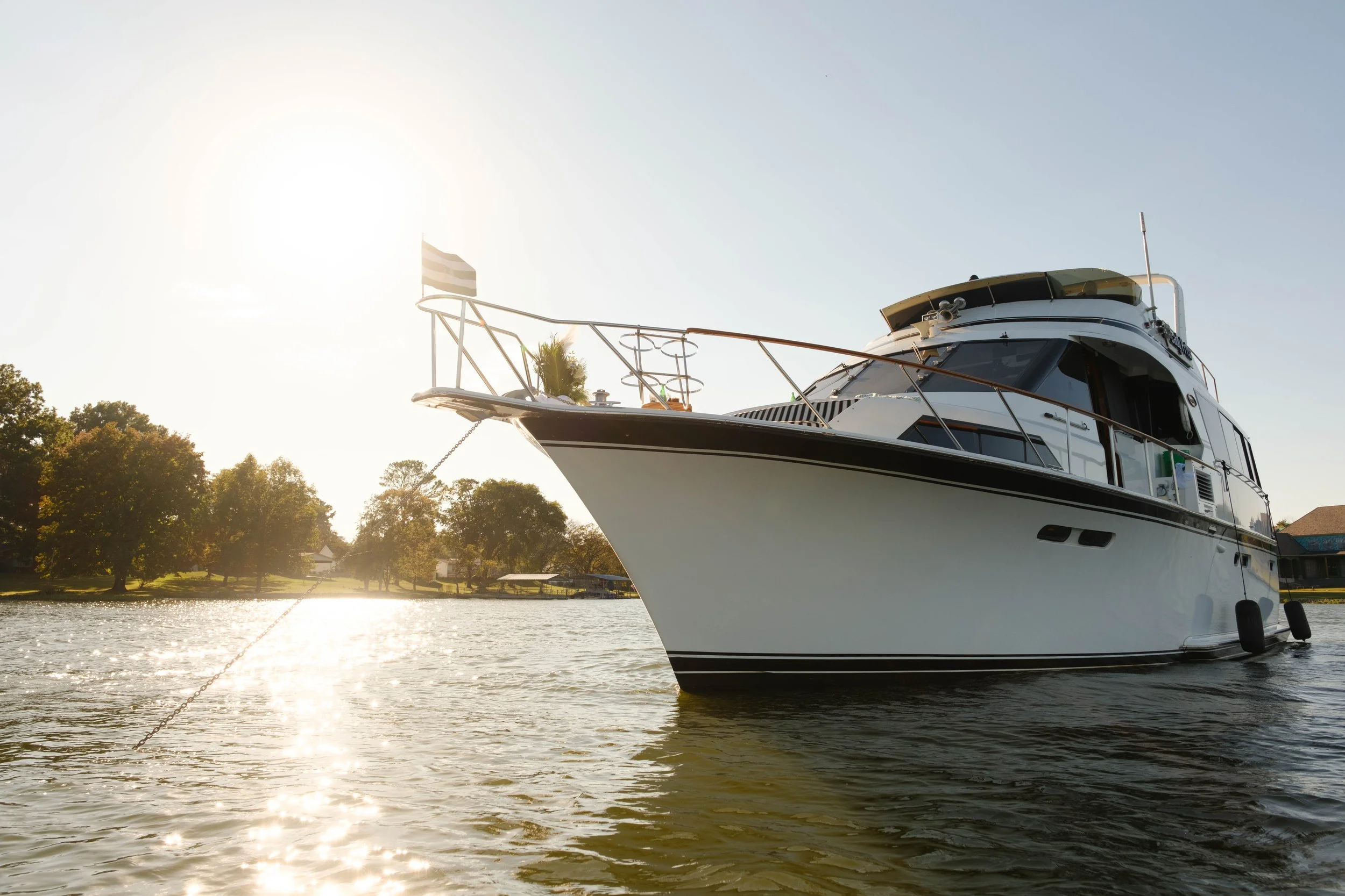 A white yacht docked on a calm river during sunset, with trees and houses in the background, and a flag flying at the bow.