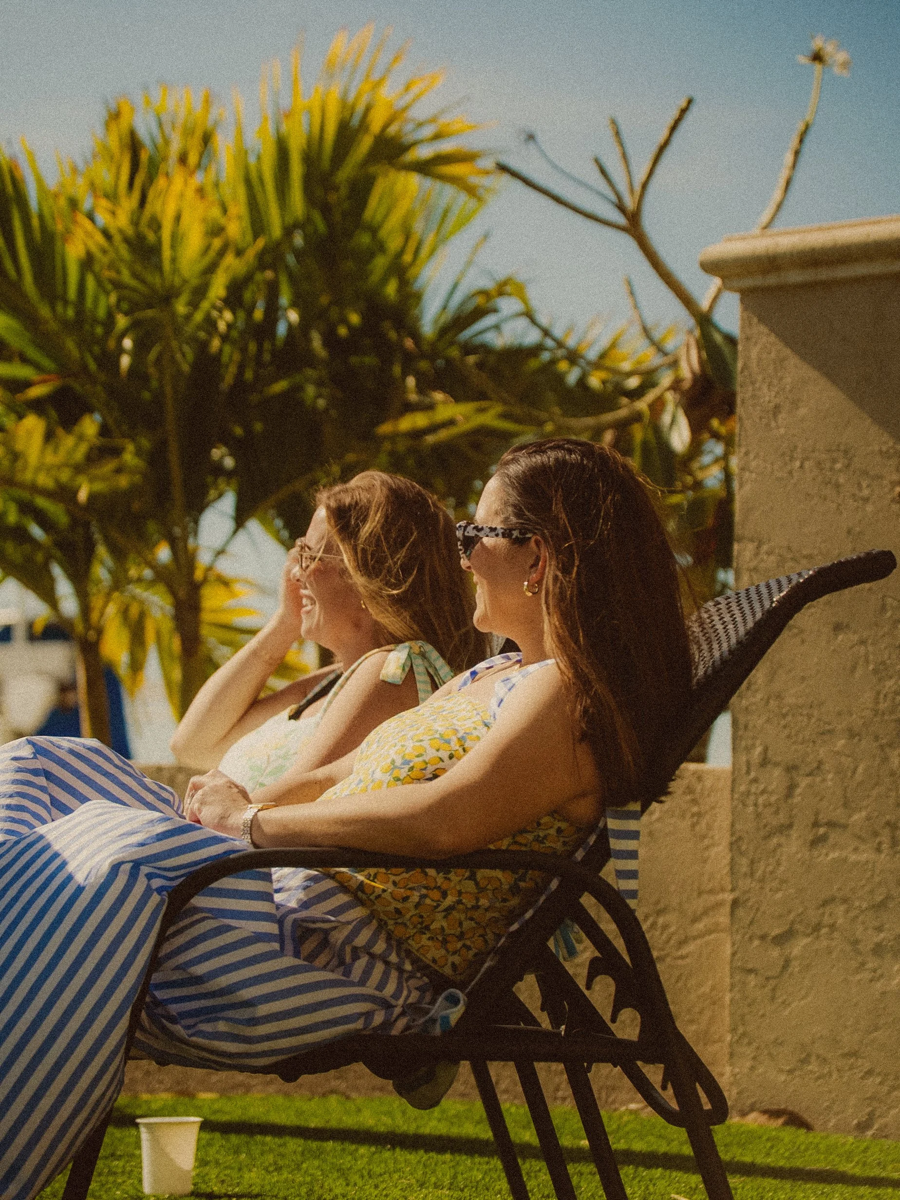 Two women with long hair and glasses are sitting outdoors on a bench, engaging in conversation. Palm trees and a building are visible in the background, suggesting a warm, tropical location.