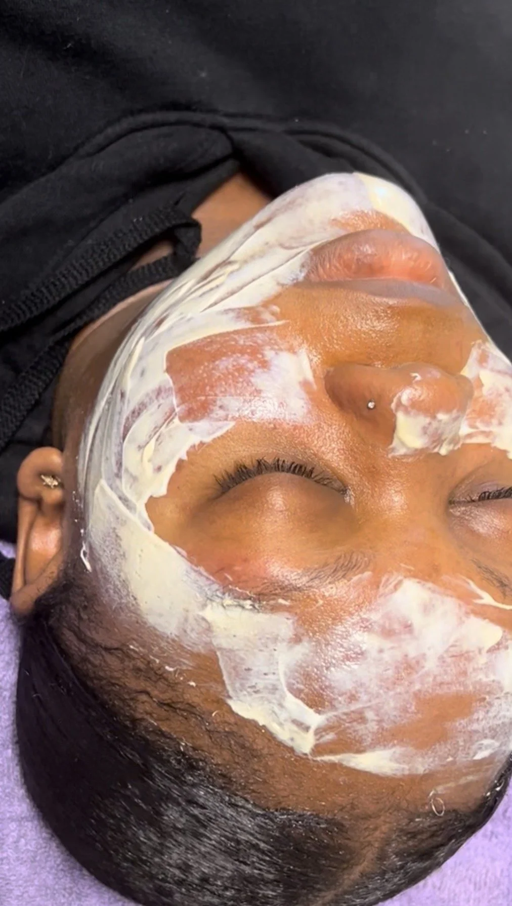 A woman lying with her eyes closed, receiving a facial treatment. Her face is covered with a mixture of white cream and orange-colored mask, possibly a skincare or beauty treatment.