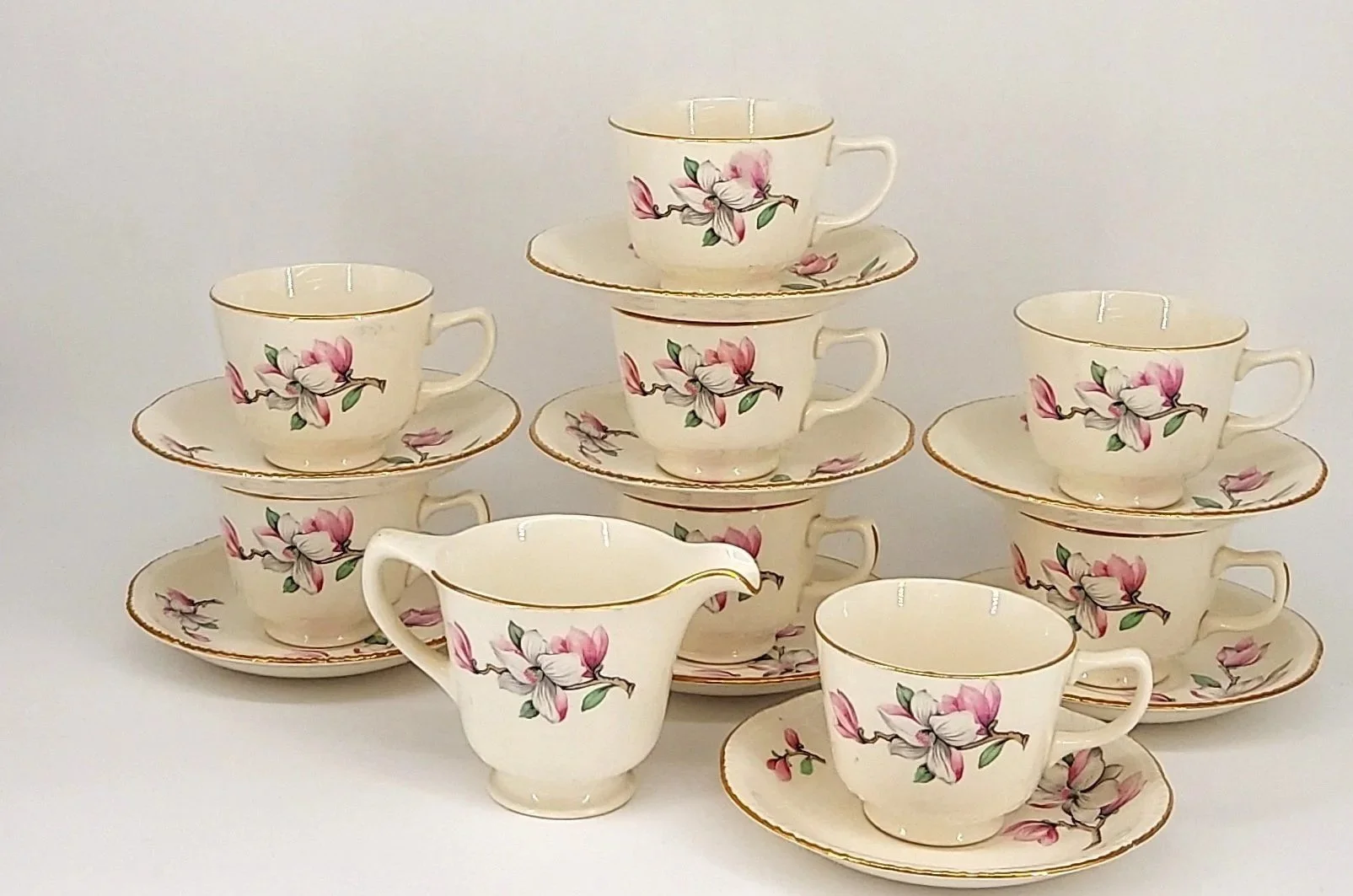 Cream-colored teacup and saucer set with pink and white flower design, with one creamer pitcher, arranged in a pyramid formation on a white background.