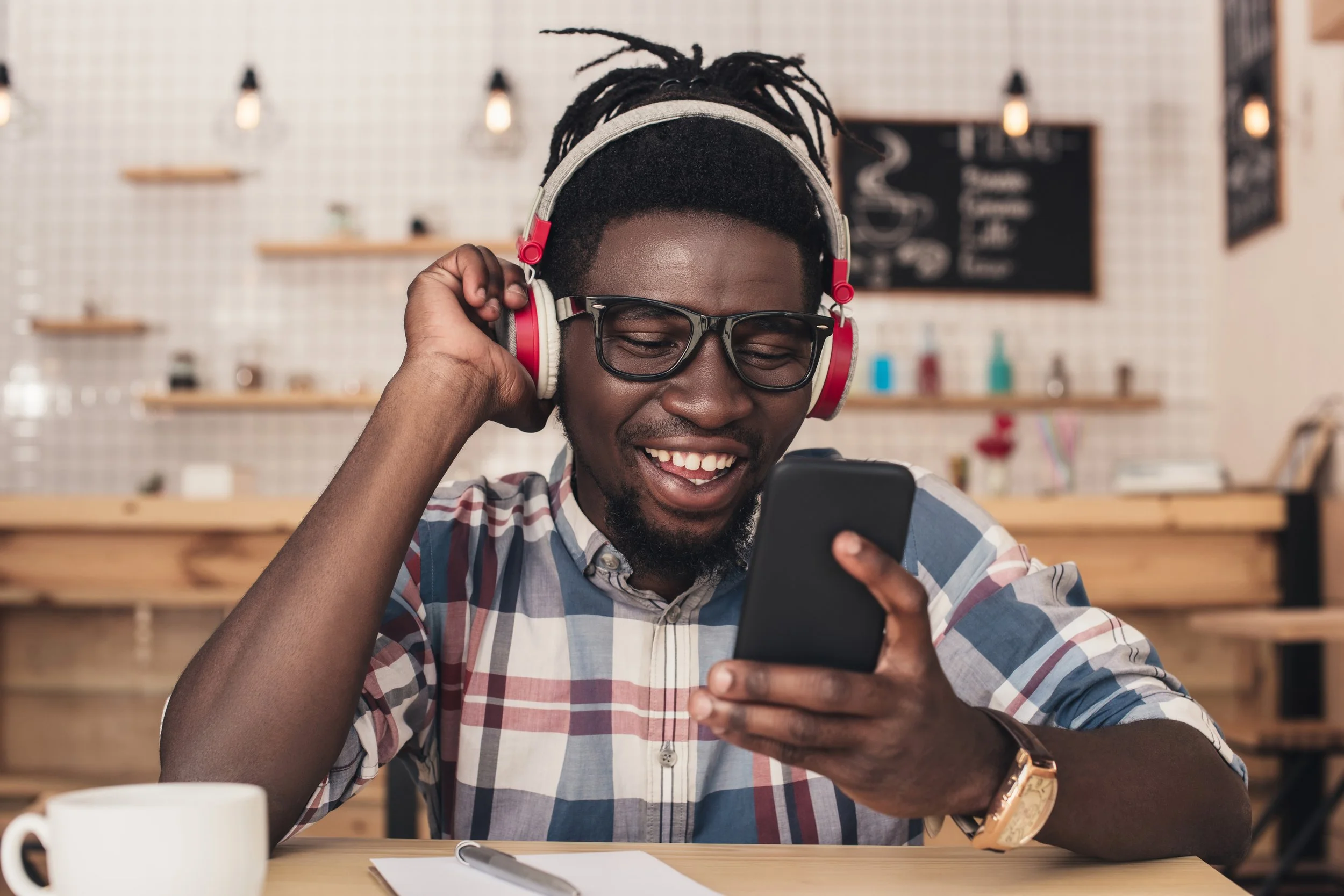 A smiling man with glasses and dreadlocks wearing headphones and a plaid shirt, looking at his phone while sitting at a table in a café.