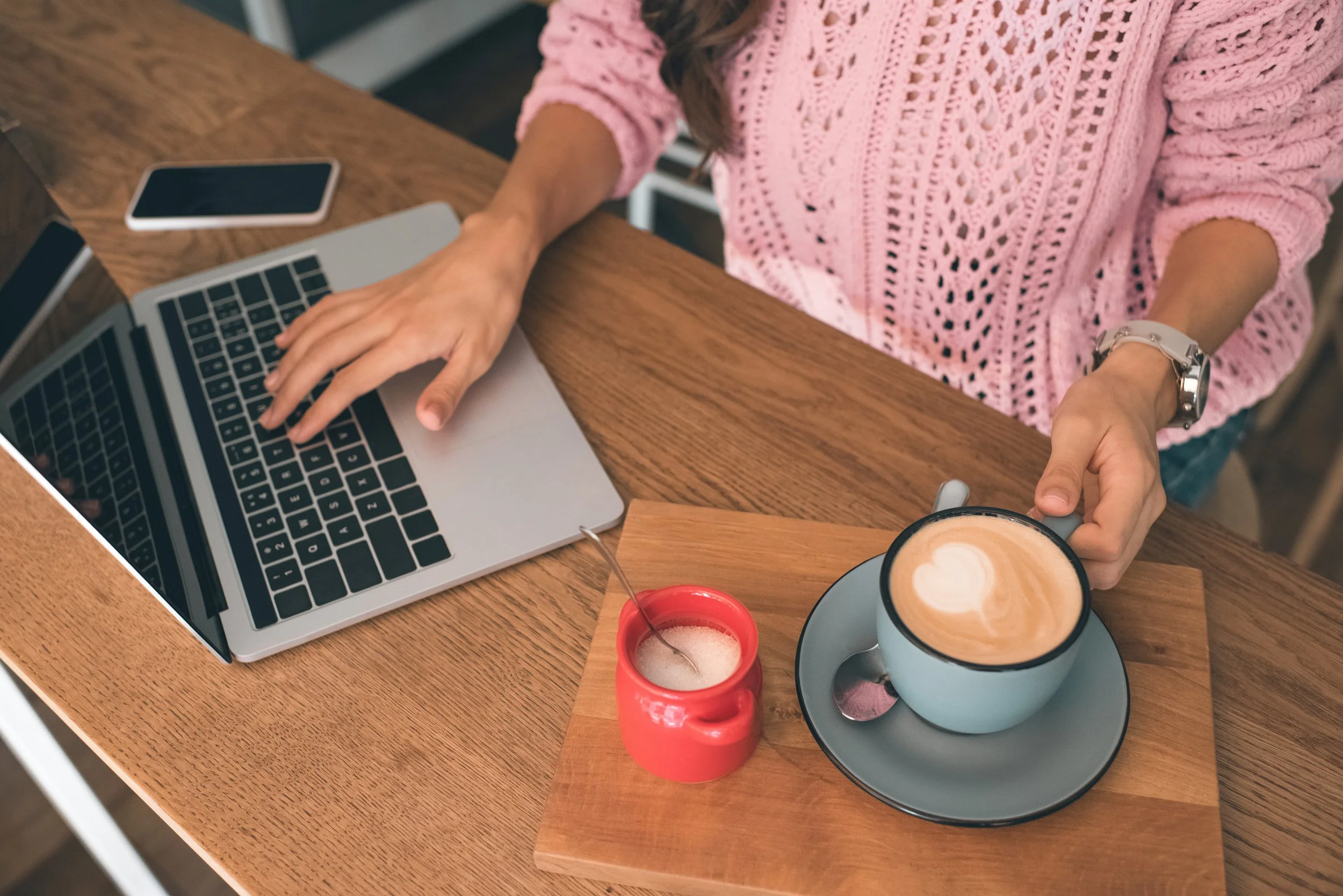 A person wearing a pink crocheted sweater is sitting at a wooden table with a silver laptop, two smartphones, a cup of coffee with foam art, a small red mortar and pestle, and a wooden spoon. The person's right hand is reaching for the coffee mug, and they are wearing a wristwatch.