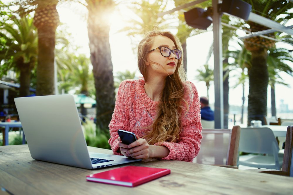 A young woman with long hair and glasses sitting outdoors at a table with a laptop and a red notebook, holding a smartphone, surrounded by palm trees and other outdoor furniture.
