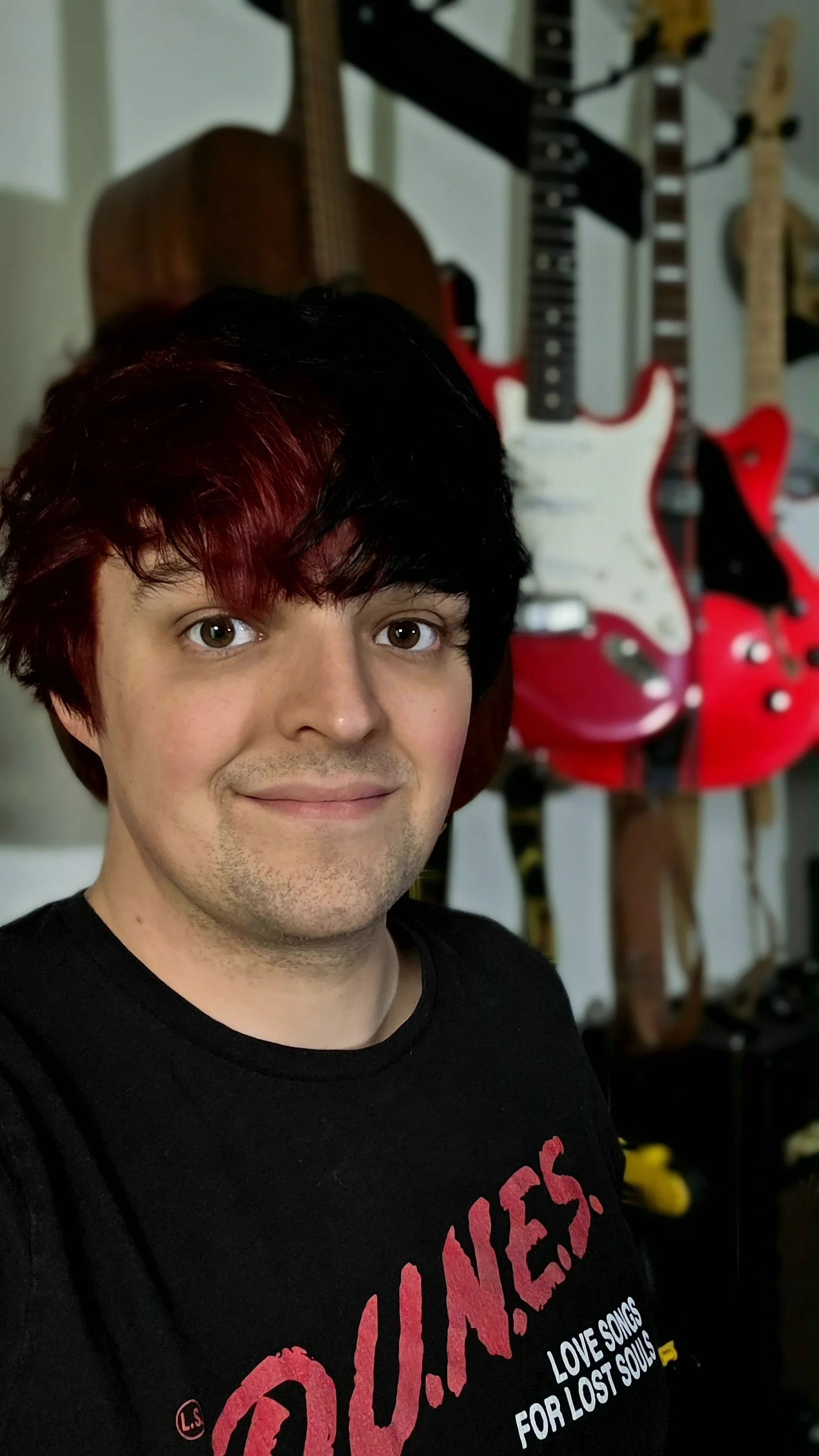 A man with short black and red hair smiling, wearing a black T-shirt with red and white text. In the background, there are multiple guitars hanging on a wall.