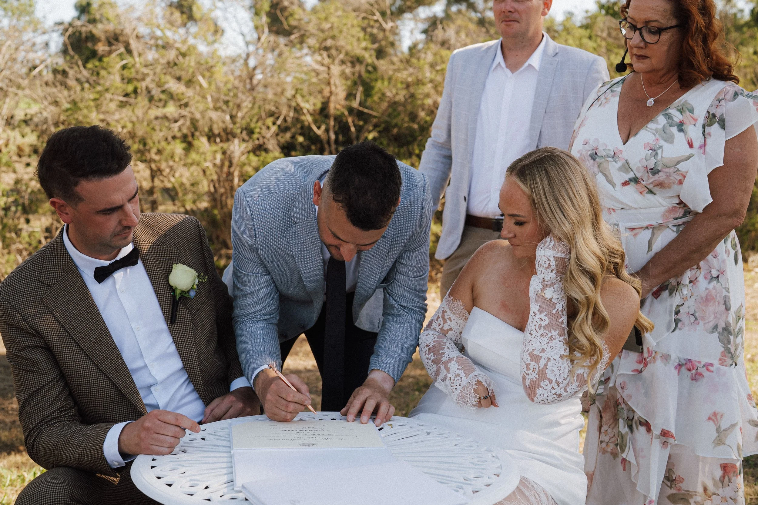 A wedding ceremony outdoors with a bride and groom signing a document, surrounded by their families and friends.