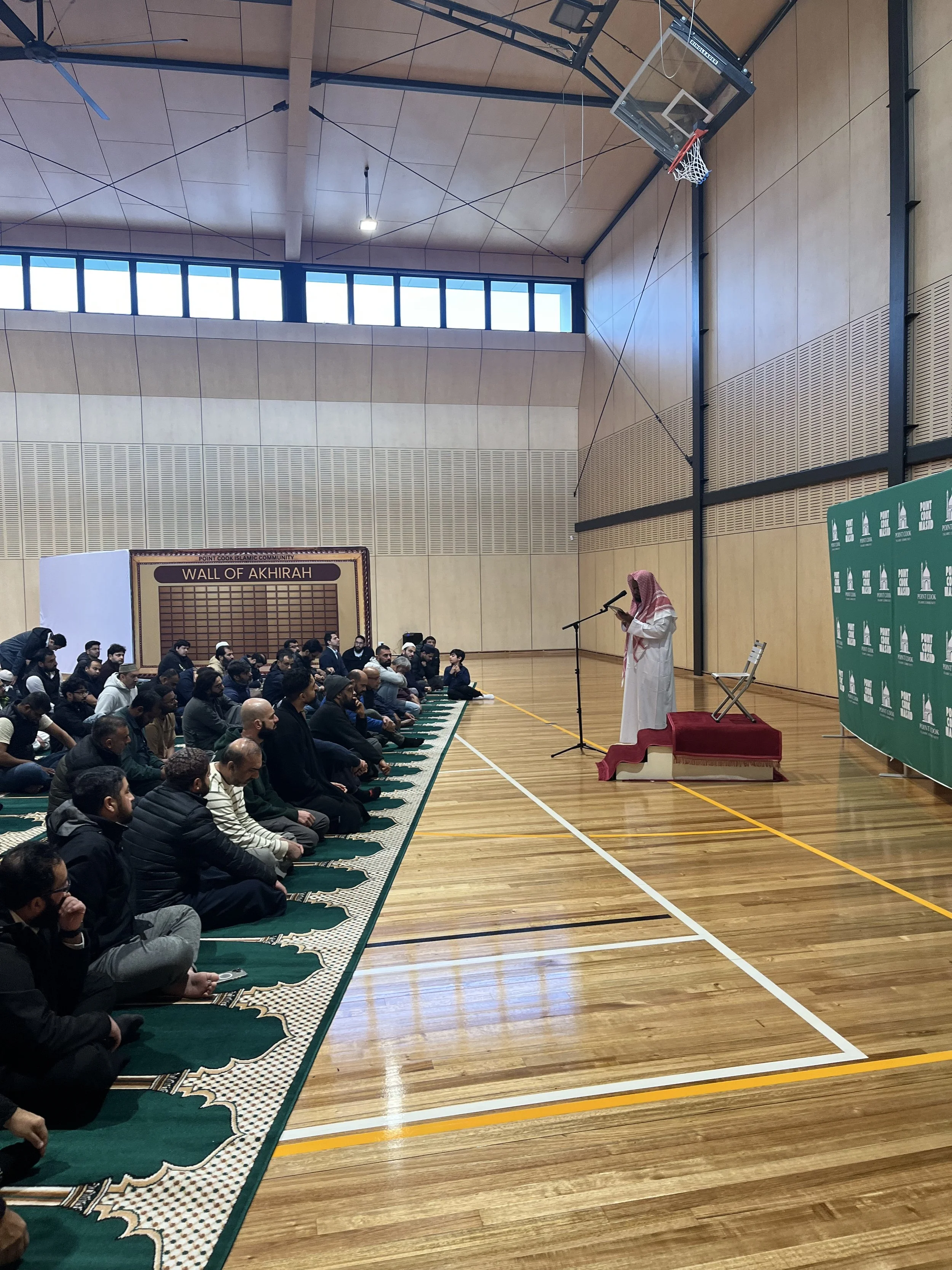 A man dressed in traditional Middle Eastern clothing stands at a microphone on a stage, addressing a seated audience of men in a gymnasium with wooden floors and high ceilings. The gymnasium has large windows and a basketball hoop.