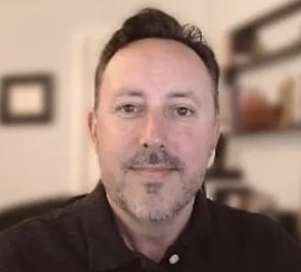 An attractive man with short dark hair and facial hair, wearing a collared black shirt, smiling slightly in an office. GIGANTIC's Principal, Marketing Executive Jason Baxter shown.