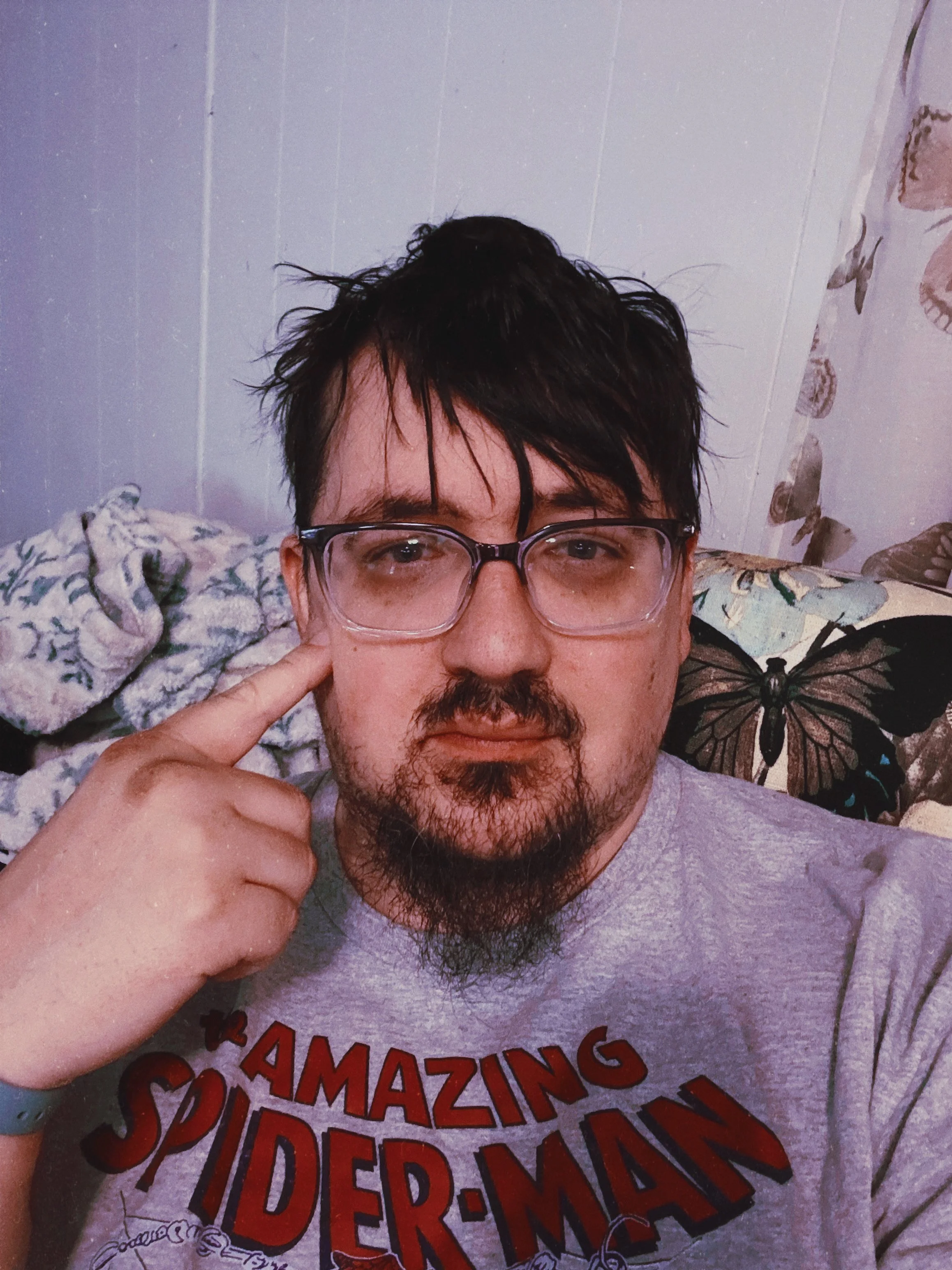 A person with dark, slightly messy hair and glasses, pointing to their temple, wearing a gray T-shirt with red text that reads "AMAZING SPIDER-MAN". Behind him is a patterned pillow with a butterfly design and a floral blanket.
