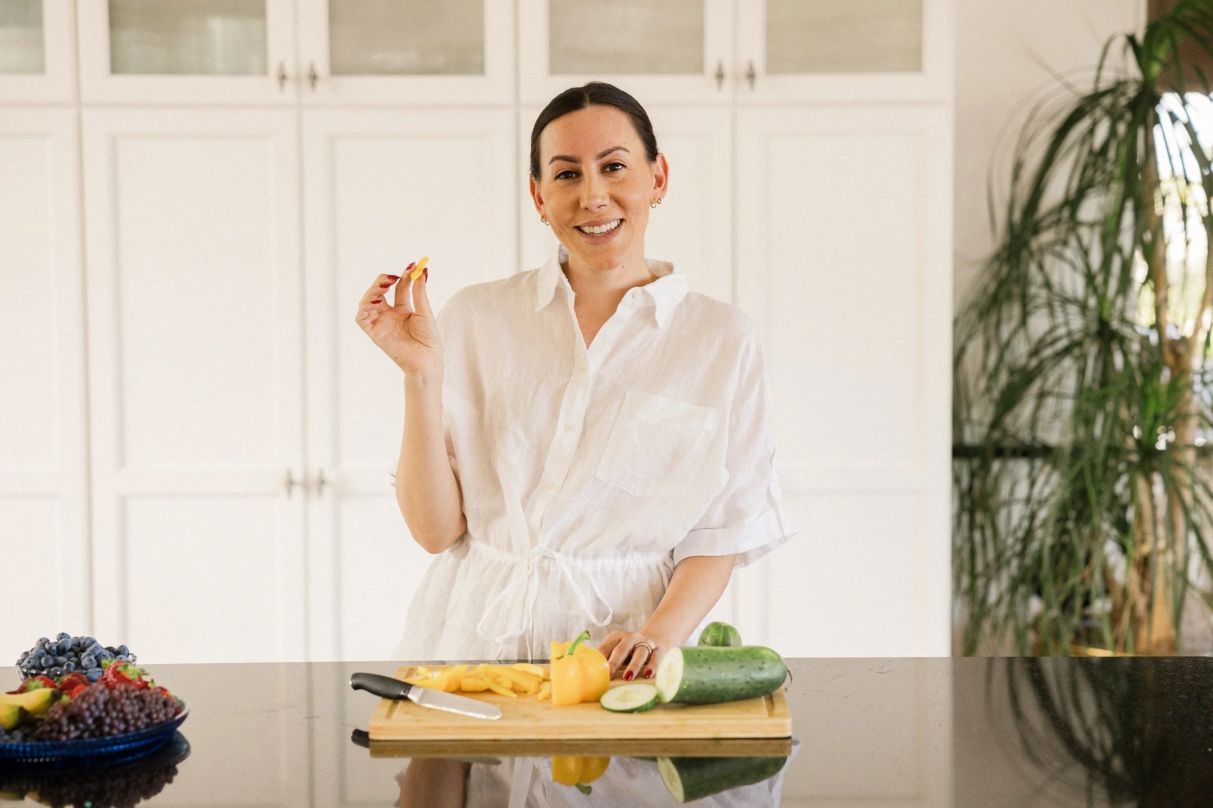 A woman in a white dress prepared to slice fruit in a bright kitchen.