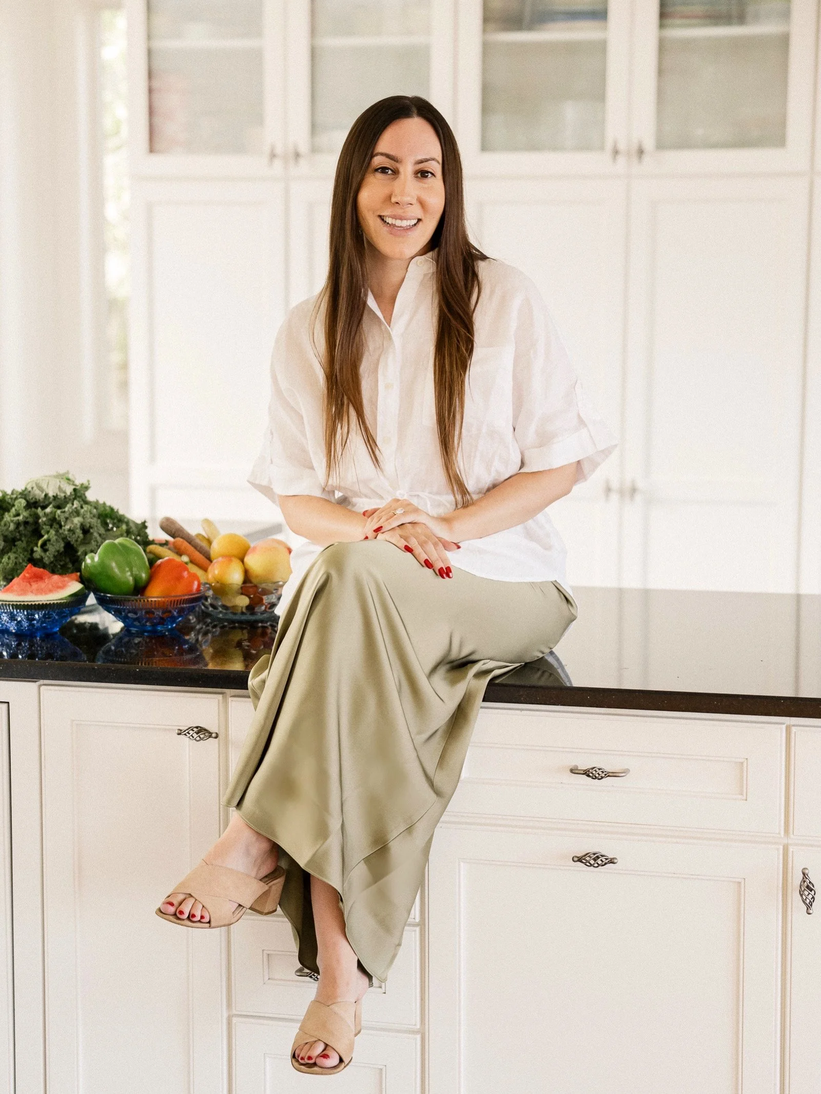 A woman with long dark hair and a white shirt sitting on a kitchen counter with colorful fruits and vegetables around her.