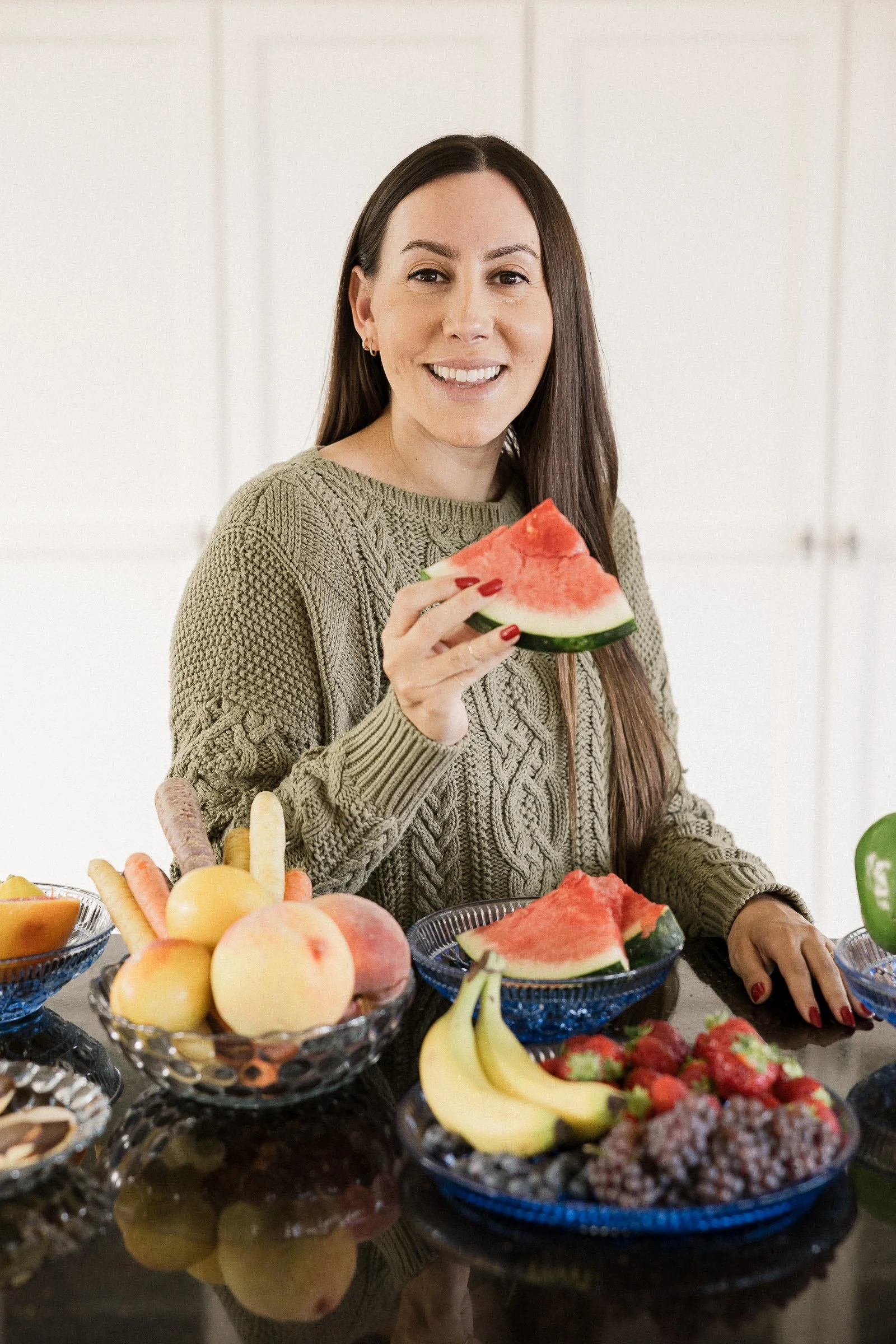 A woman holding a slice of watermelon standing at a table filled with various fruits including bananas, peaches, strawberries, and apples, with a white kitchen background.
